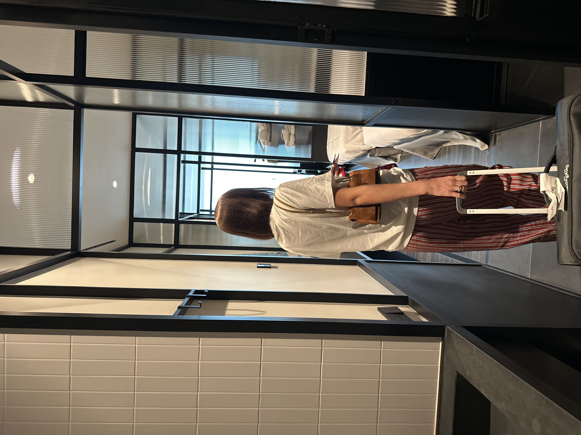 A person stands in an interior space with a suitcase. The interior features white tiled walls, black framing elements, and vertically textured glass panels. The person has brown hair and wears a white shirt and striped pants, carrying a brown purse. A room with a bed is visible through an opening. The location is suggested to be in Tbilisi, Georgia.