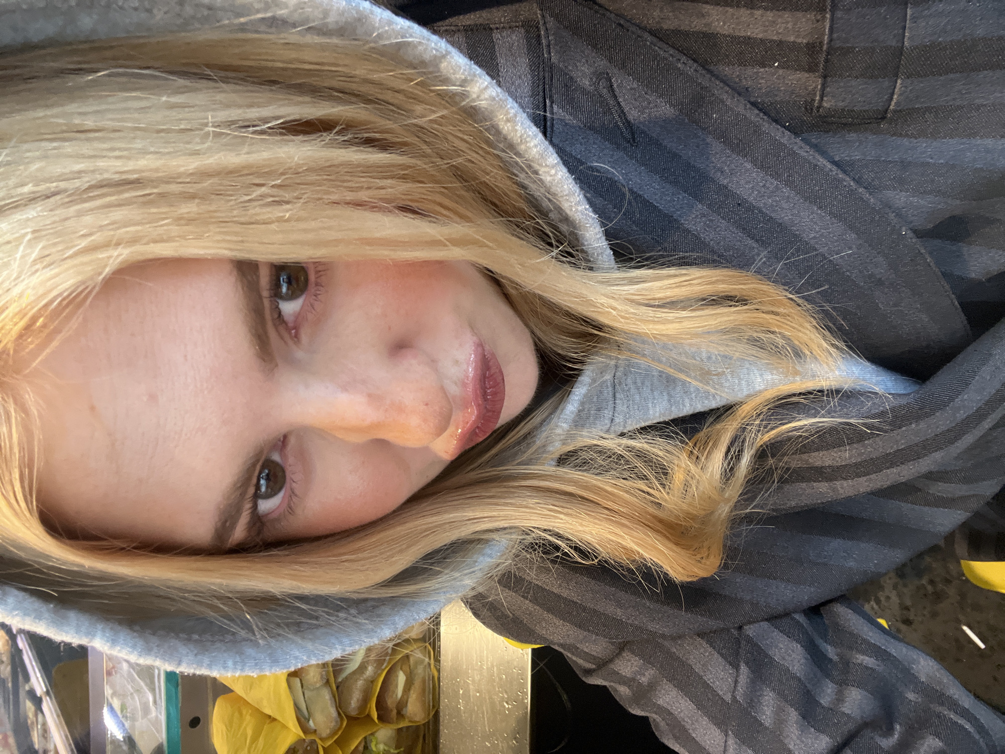 A light-skinned person with blonde hair and brown eyes, wearing a hooded gray sweatshirt and a dark gray striped blazer is visible. A clear plastic container with yellow contents is visible on the left side of the image. The location in the city of Paris, France, is not discernible.