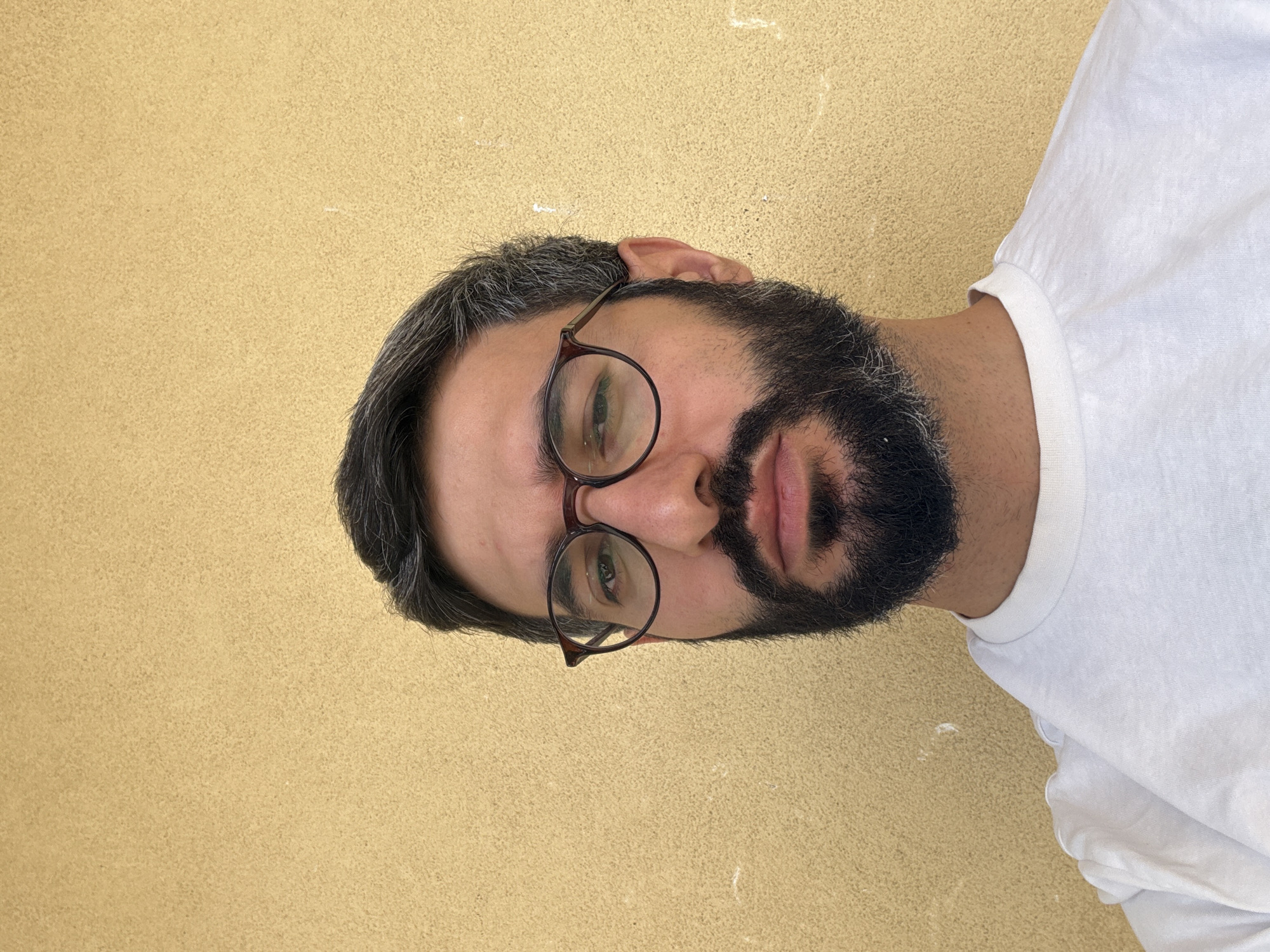 A man wearing glasses and a white t-shirt stands against a textured yellow wall. His face is turned towards the camera, and he has a beard. The image appears to be a selfie, taken outdoors. There are no discernible landmarks that would indicate a specific location in Milan.
