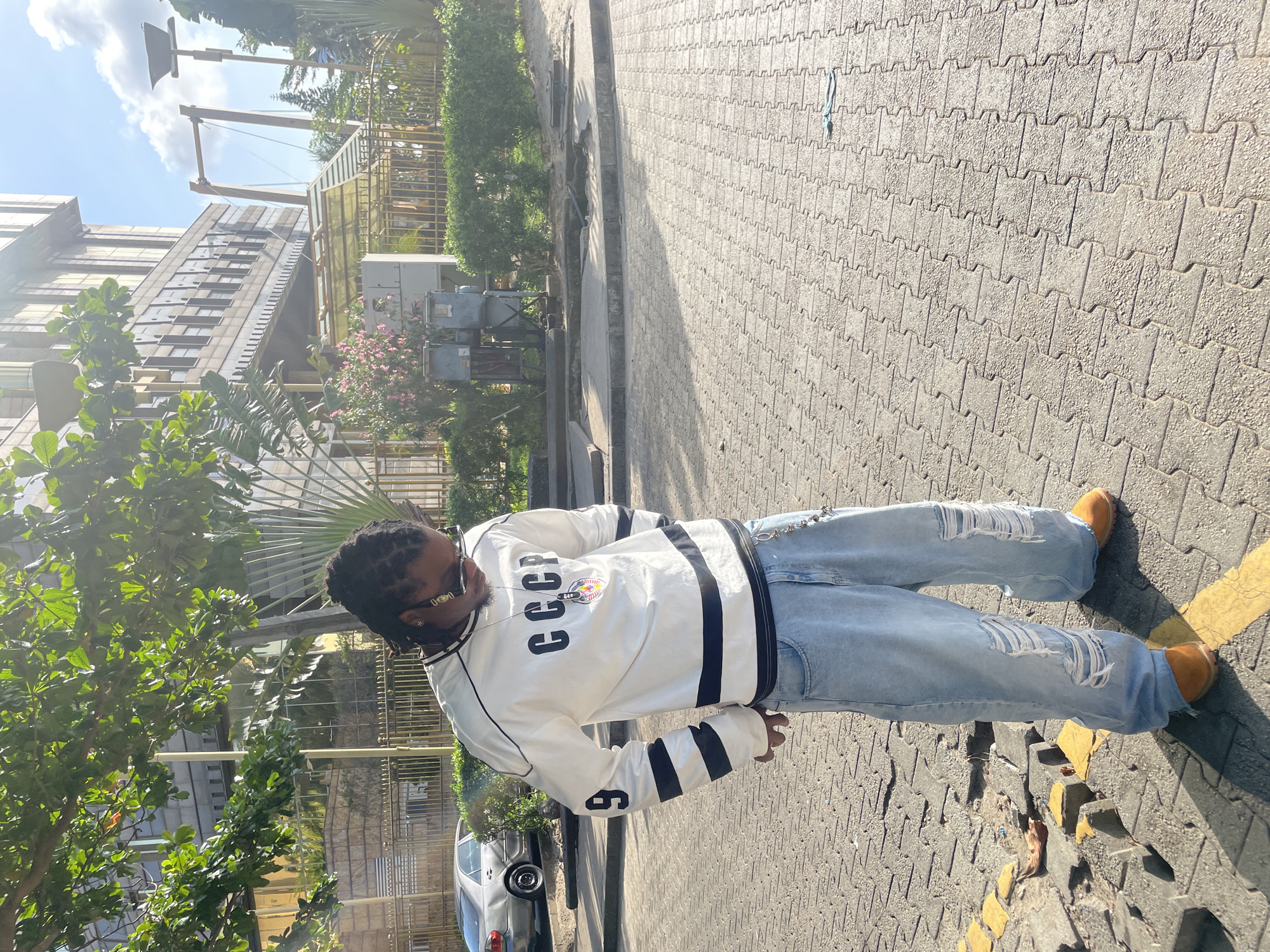A person stands on a textured sidewalk in an urban setting. The person is wearing a white jacket with black stripes and the letters "CCCP", ripped blue jeans, and brown shoes. Buildings and trees are visible in the background, along with a car parked on the side of the road. The sidewalk is composed of interlocking pavement stones and has yellow painted lines.
