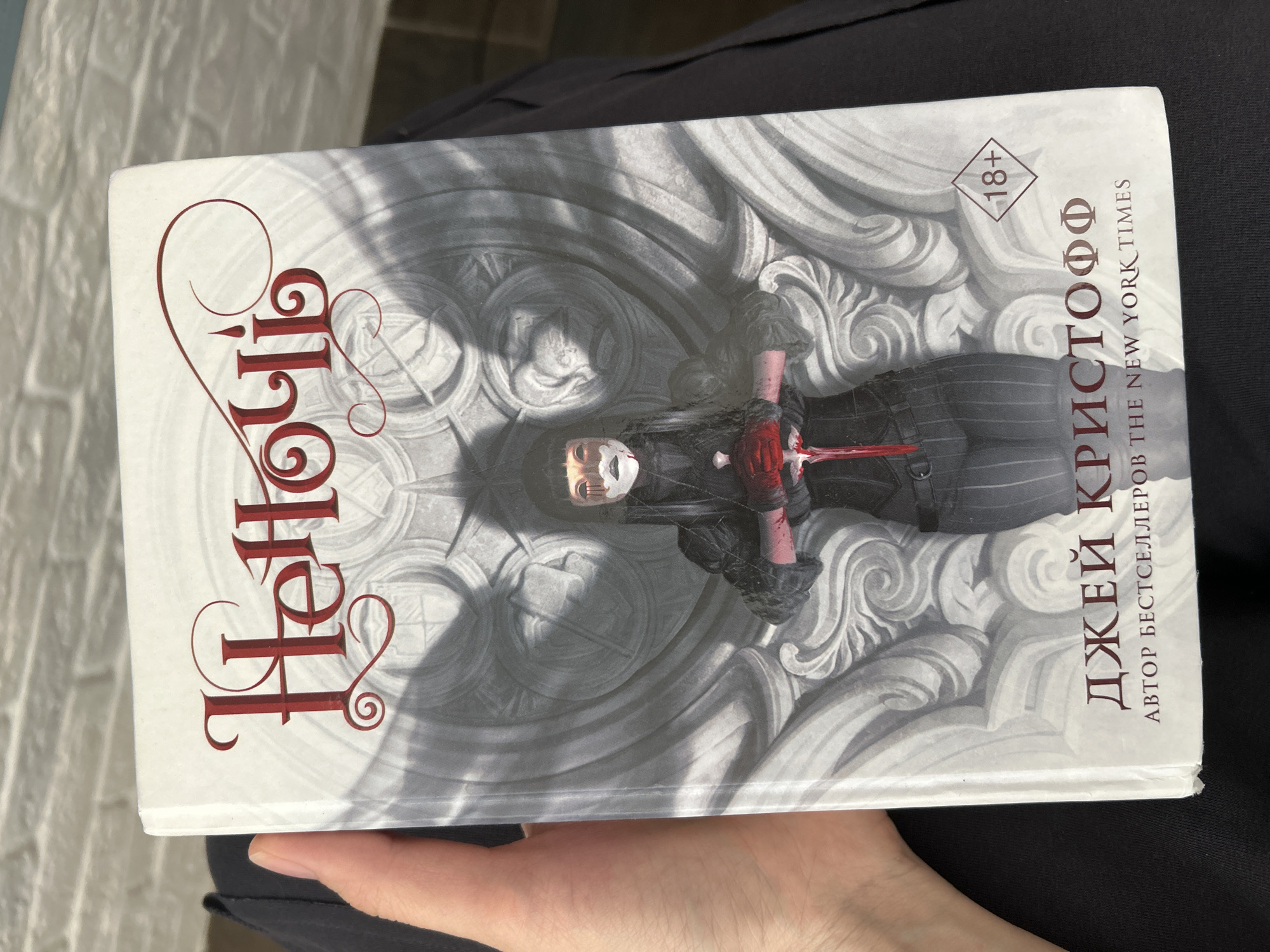 The media file shows the cover of a book, held in a person's hand. The book cover features an illustration of a person with a mask and red gloves, holding a bloody dagger. The background of the illustration is a stone carving with intricate patterns.
Visible text on the cover includes:
- Cyrillic text: "ДЖЕЙ КРИСТОФФ" (likely the author's name)
- "АВТОР БЕСТСЕЛЛЕРОВ THE NEW YORK TIMES" (author of bestsellers The New York Times)
- "18+" inside a diamond shape.
- Stylized red Cyrillic letters spelling out "Hellion" or a similar word, with ornate swirls.
The lighting suggests it is daytime, with sunlight casting shadows on the book cover. The book appears to be oriented vertically in the image. The person's hand holding the book is visible at the bottom. The overall aesthetic of the cover is dark and dramatic, hinting at a fantasy or horror genre.