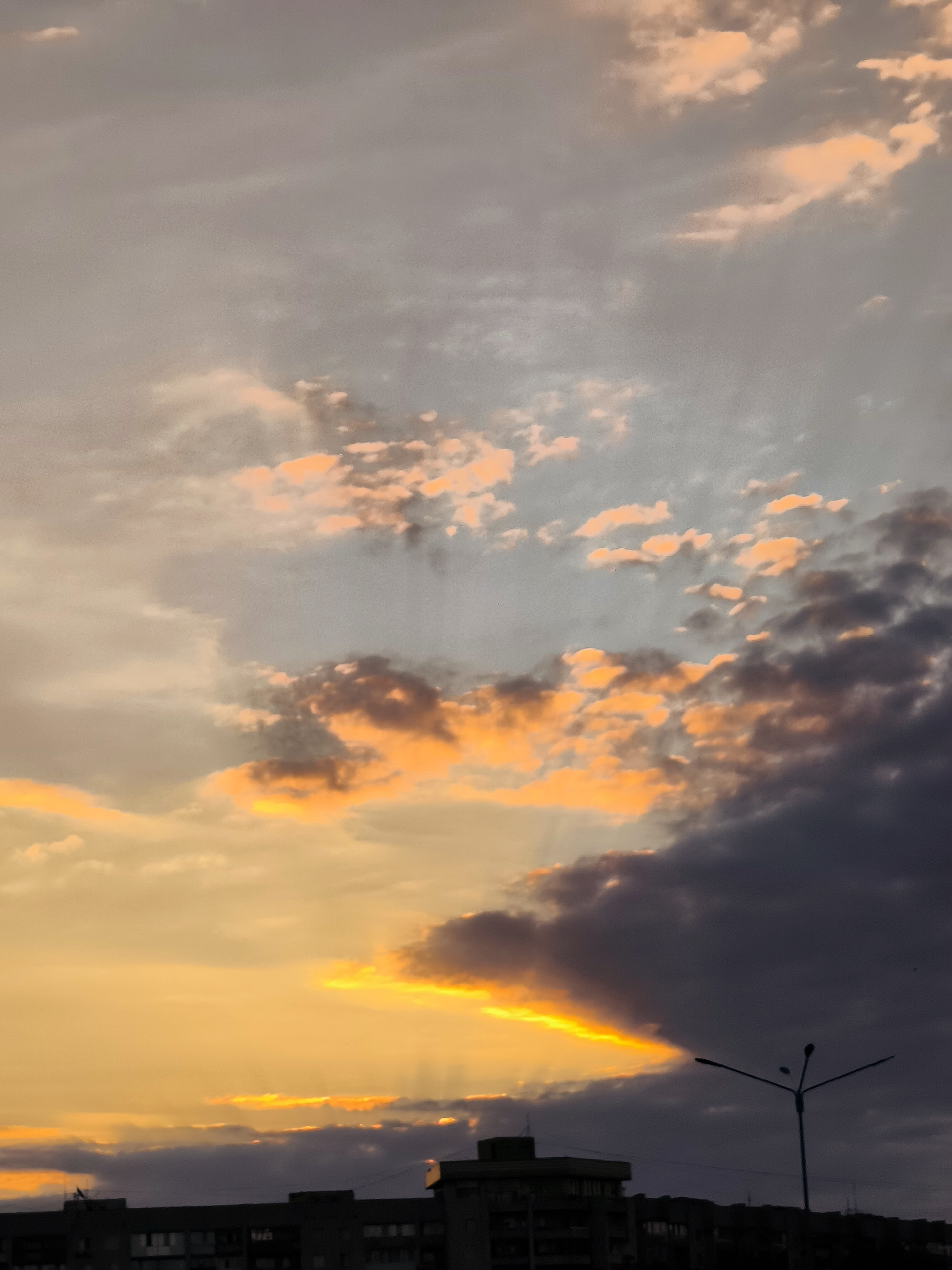 The image captures a sunset over the city administration area of Qarağandı, Kazakhstan. The sky is a dramatic mix of colors, with bright orange and yellow hues from the setting sun illuminating the edges of dark, gray clouds. Streaks of light pierce through gaps in the clouds, creating a dramatic effect. In the foreground, silhouetted against the vibrant sky, are the rooftops of apartment buildings and a street lamp. The overall scene is one of a vibrant, natural phenomenon occurring over an urban landscape.
