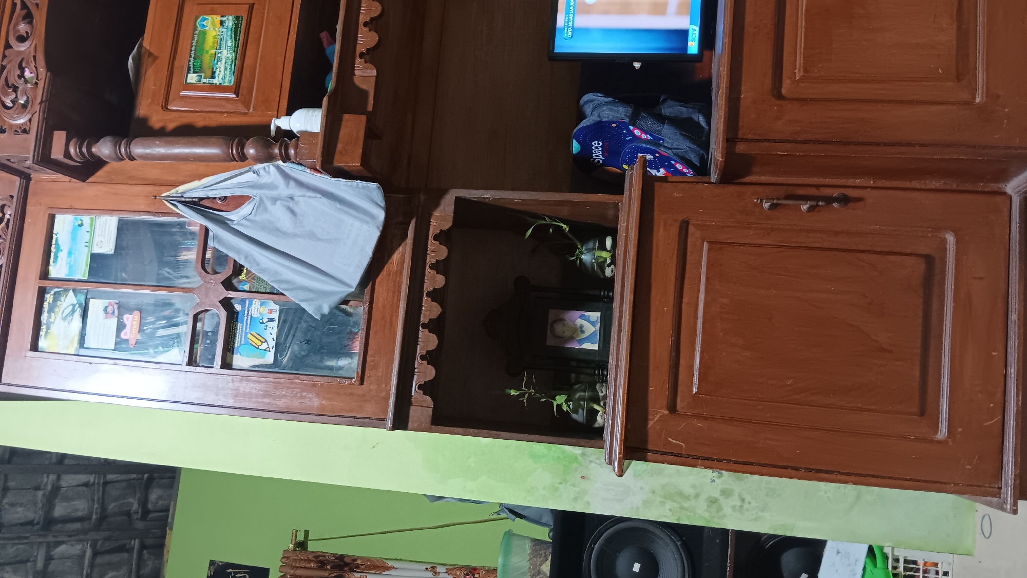 The image shows a wooden cabinet in a room. The cabinet has multiple shelves and drawers. On one of the shelves, there is a framed photograph of a child, two small potted plants in glass vases, and a gray tote bag hanging from a hook. Above this shelf, there is a glass-doored compartment with various items displayed inside. To the right of the cabinet, a television screen is visible, displaying what appears to be a broadcast with the SCTV logo at the bottom. The background is a green wall and a part of a tiled ceiling. On the left side of the image, a large speaker is partially visible. The overall scene depicts an indoor setting, likely a living room or a study, in Sonobekel, Indonesia.