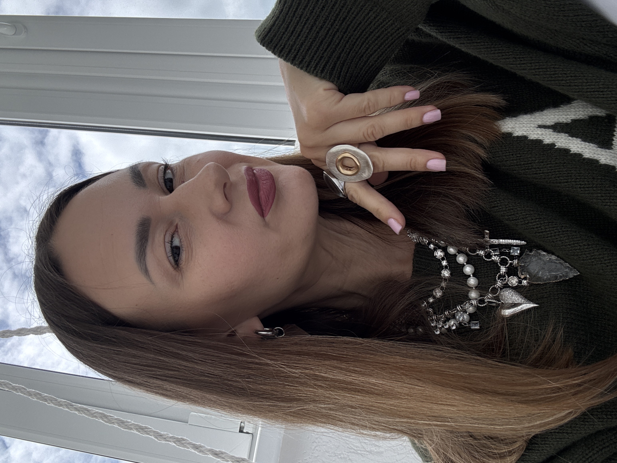 A young, fair-skinned woman is pictured indoors, potentially near a window, as suggested by the diffused daylight and the hint of the outside sky. She wears a dark green sweater with a light-colored pattern at the top. Her hair is long, straight, and light brown. She is adorned with jewelry, including a large, oval ring on her finger and a necklace featuring pearls, decorative metal pieces, and a dark, arrowhead-shaped pendant. Her fingernails are painted a light pink. There is no explicit evidence that this image was taken in Sochi, Russia.