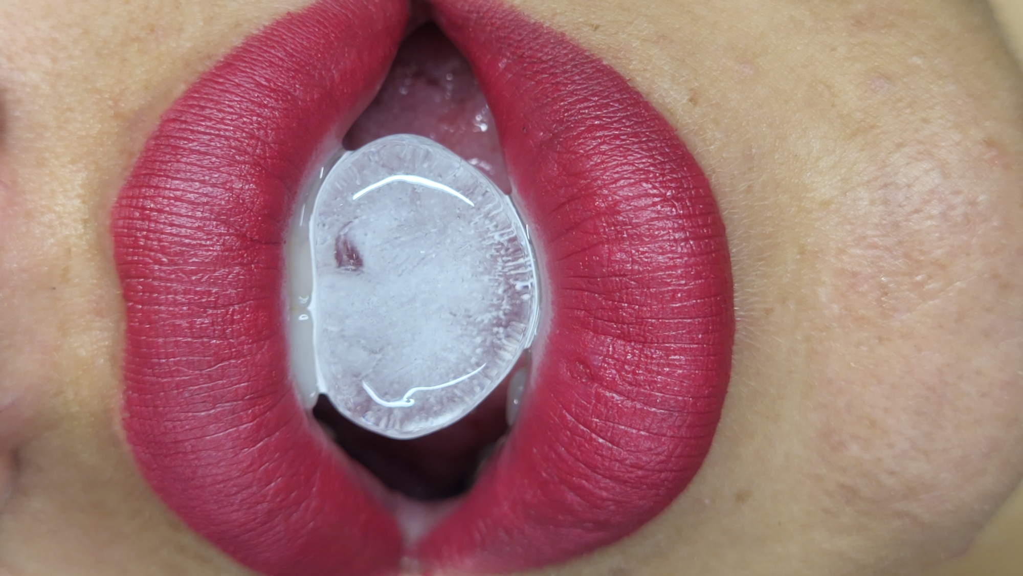 A close-up, eye-level shot shows a person's mouth with bright red lipstick and an ice cube held between their lips. The ice cube appears somewhat melted and is partially obscuring the tongue. The lighting is bright and even, highlighting the texture of the lips and the detail of the ice. The background is out of focus, suggesting a shallow depth of field. The overall impression is one of sensuality and coolness. The image does not contain any elements that would indicate a location in Horishni Plavni, Ukraine.