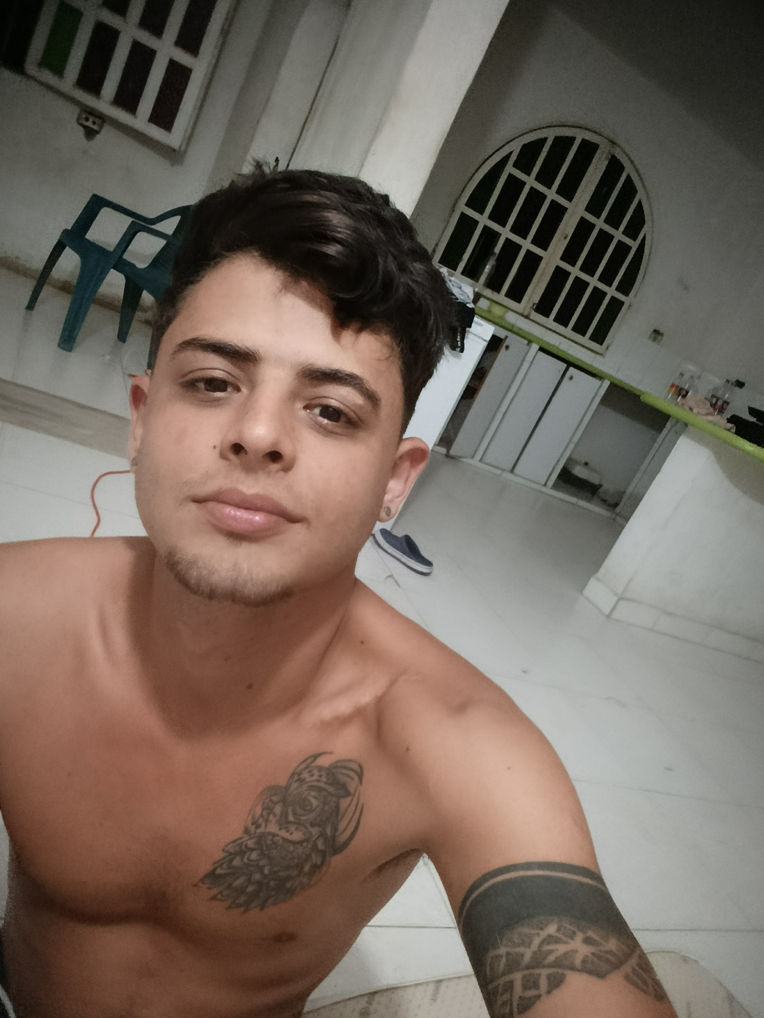A shirtless, light-skinned male with dark hair and light stubble is positioned in the foreground, taking a selfie. He has two visible tattoos: one on his upper left chest of an owl and another on his left upper arm of a geometric pattern. He appears to have an earring on his left ear. Behind him, in a sparsely furnished room, a light blue plastic chair is visible in the upper left corner. In the background, there is an arched window with grid-like panes. Also visible is a white kitchen counter and cabinets, along with a shoe near the base of the cabinet. The location is in the city of Unknown, Unknown. The floors are white tile.