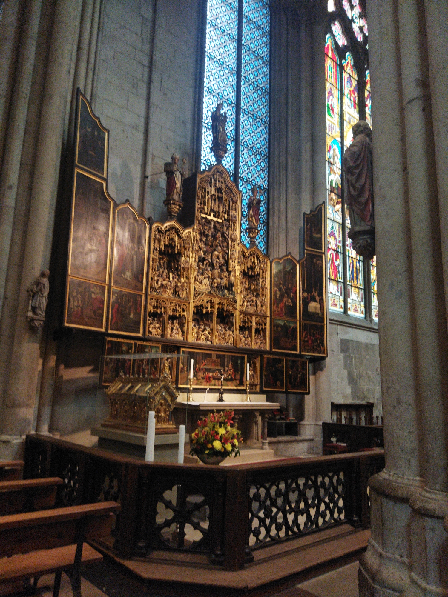 The image shows the interior of a church, likely St. Gereon in Monheim am Rhein, Germany, judging by the style of the architecture and the presence of religious artifacts. The focal point is an elaborate altar with a large, gilded retable featuring numerous carved figures in various scenes, possibly biblical. To the left of the retable, there are several framed paintings arranged in a triptych-like formation, also depicting religious imagery. Above the altar, a large stained-glass window with intricate geometric patterns and some figural elements casts colored light into the space. To the right of the altar, a tall, slender column frames a section of a colorful stained-glass window with prominent red and blue hues. A statue of a robed figure, likely a saint, is visible near the column. In the foreground, a dark wooden railing with ornate carvings separates the viewer's space from the altar area. The floor appears to be tiled or paved. The overall atmosphere is solemn and historical, indicative of a place of worship. There are no people visible in the image.