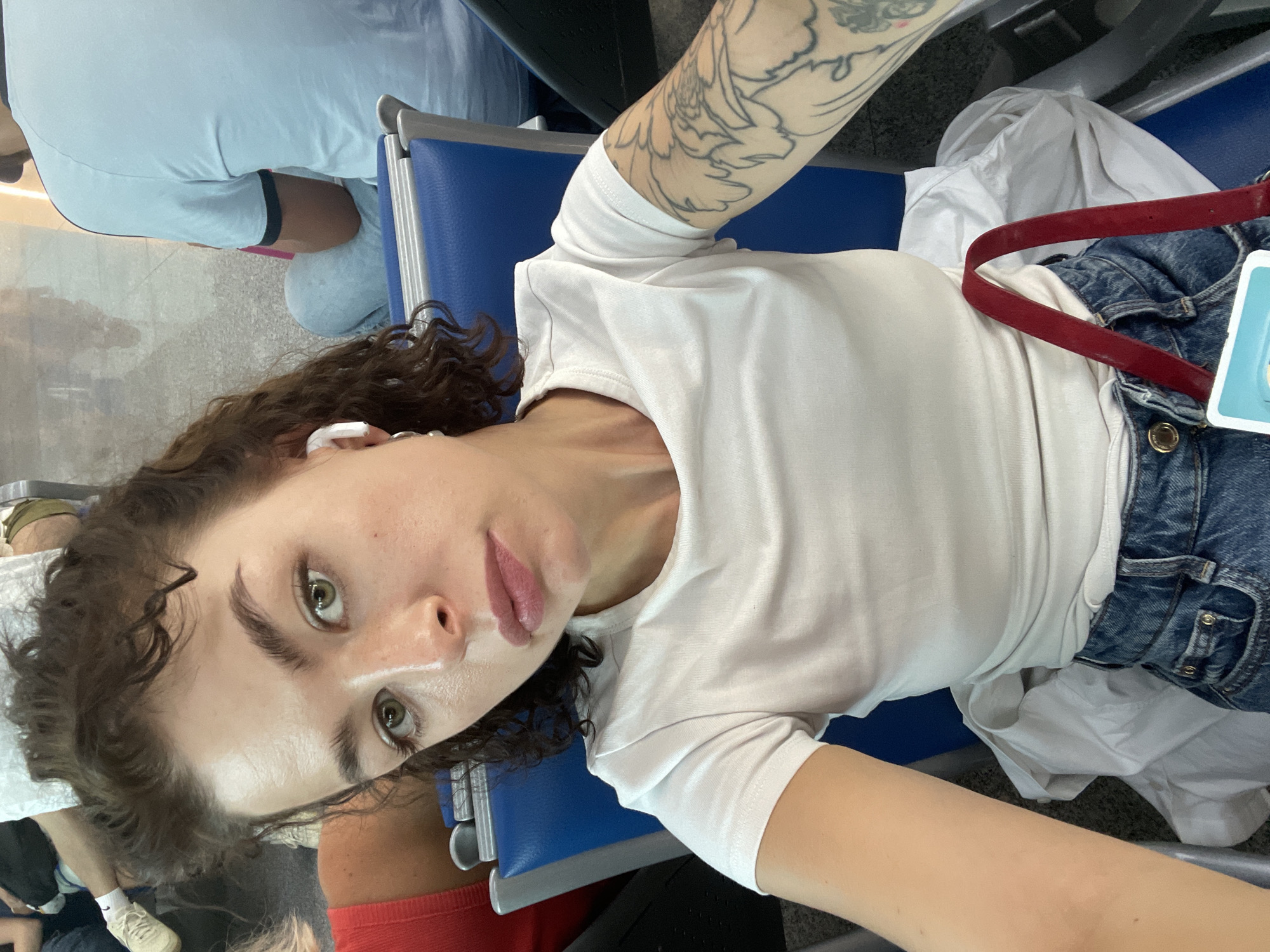A woman with curly brown hair and green eyes takes a selfie while sitting in a blue airport chair. She is wearing a white t-shirt, blue jeans, and white earbuds. Her left arm, visible on the right side of the frame, has a large floral tattoo. A red strap from a bag is draped over her lap. The background is blurred, but other people and airport seating can be discerned. The specific location within Podgorica, Montenegro is not determinable from the visual information provided.