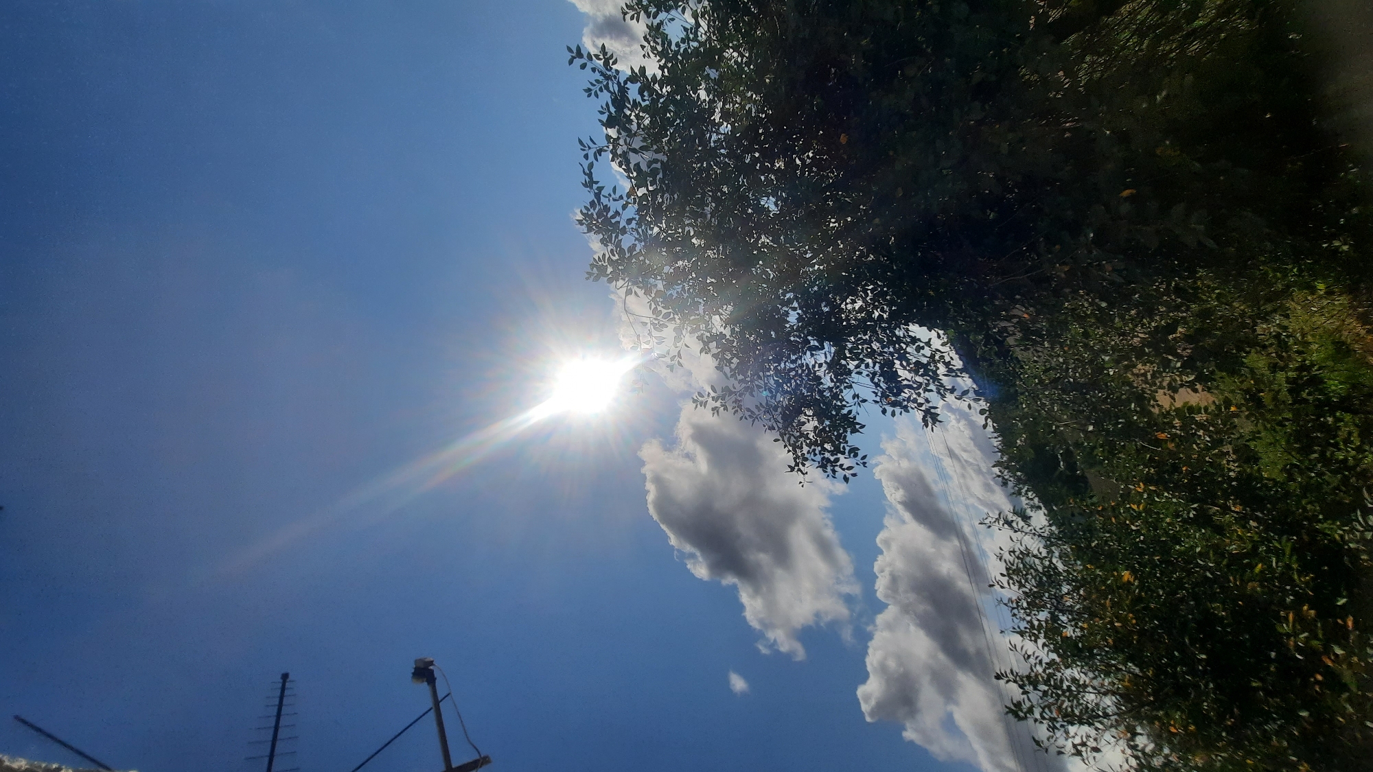 The image depicts a bright, sunny day with a clear blue sky. The sun is prominent, casting a strong glare and sunbeams that extend across the upper portion of the frame. Several white and gray clouds are scattered across the sky. In the foreground, the upper branches and leaves of a deciduous tree are visible, partially obscuring the sun. To the left of the frame, a dark antenna or transmission tower is visible against the sky. The lower part of the image shows more foliage and a hint of a grassy or wooded hillside in the background. There are no people or animals present in the image. The scene appears to be an outdoor setting, potentially a park or a rural area. The image does not provide any specific visual cues to pinpoint its exact location within the city of Шклоў, Belarus.
