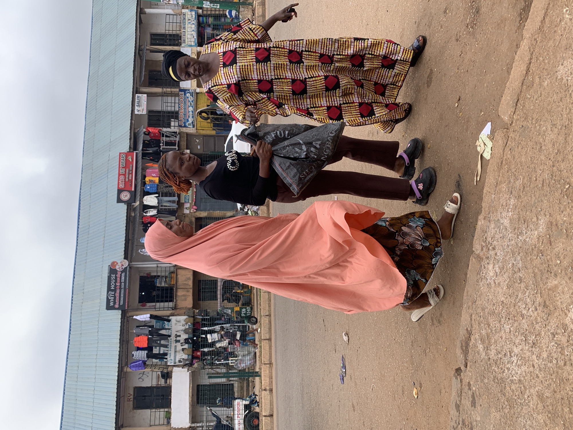 Three women are walking on the side of a street in Bukuru, Nigeria. The woman on the left is wearing a pink hijab and a patterned skirt, and she is carrying a black plastic bag. The woman in the middle is wearing a black long-sleeved shirt and brown pants, and she is also carrying a black plastic bag. The woman on the right is wearing a colorful patterned dress and a headscarf. In the background, there are shops with signs written in English. The sky is overcast, and the road is paved with asphalt. There are a few vehicles visible in the background, including a tricycle and a car. The overall scene depicts a busy street market environment.