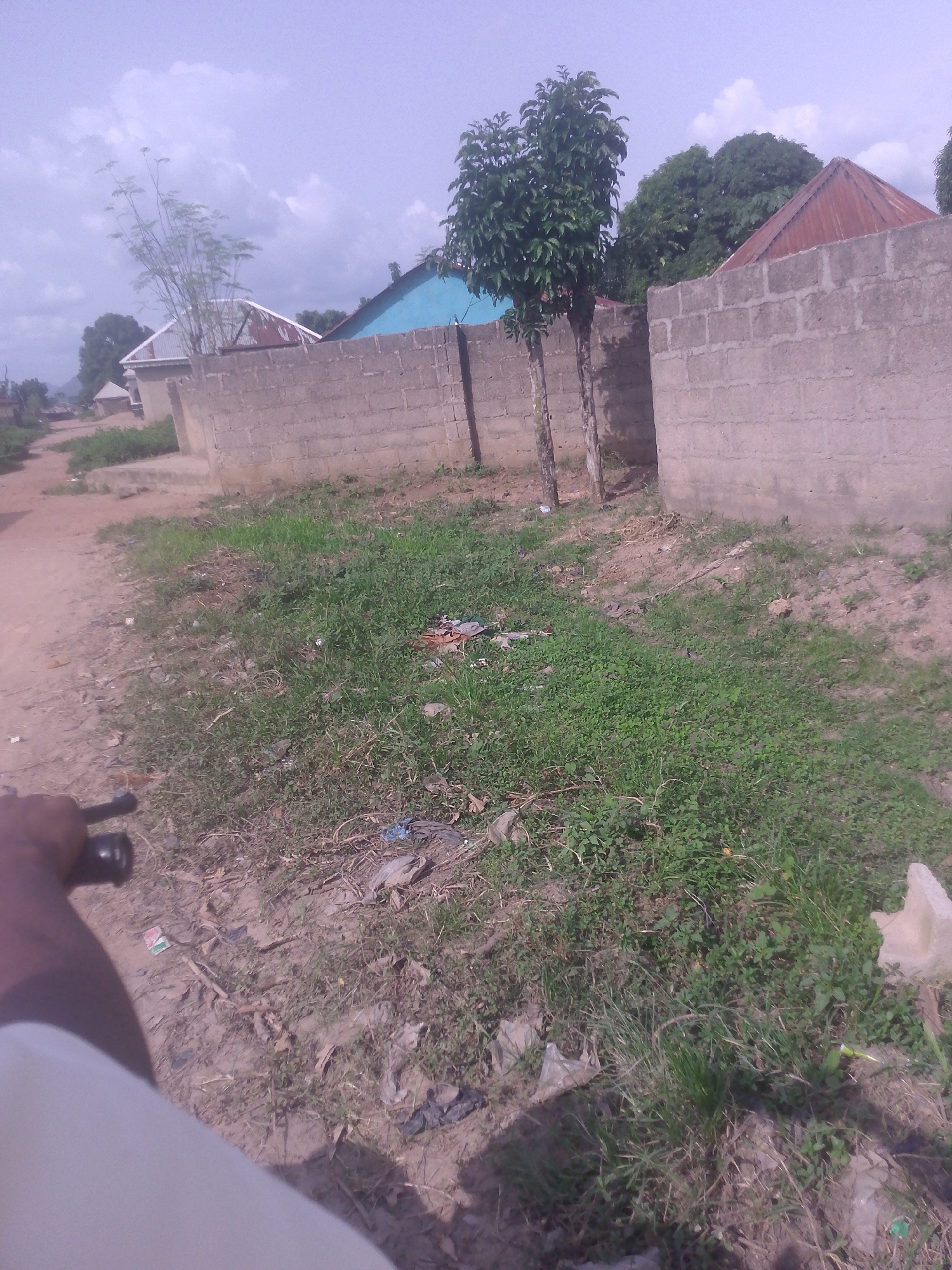 The image depicts a street scene in Serti, Nigeria. A dirt road runs along the left side of the frame, with a hand holding a black object, possibly a motorcycle handlebar, visible in the foreground. The road is bordered by overgrown grass and scattered debris, including plastic bags and other refuse. In the background, a wall constructed of concrete blocks runs along the property line. Beyond the wall, houses with corrugated metal roofs are visible, along with some trees and vegetation. The sky is overcast with clouds.