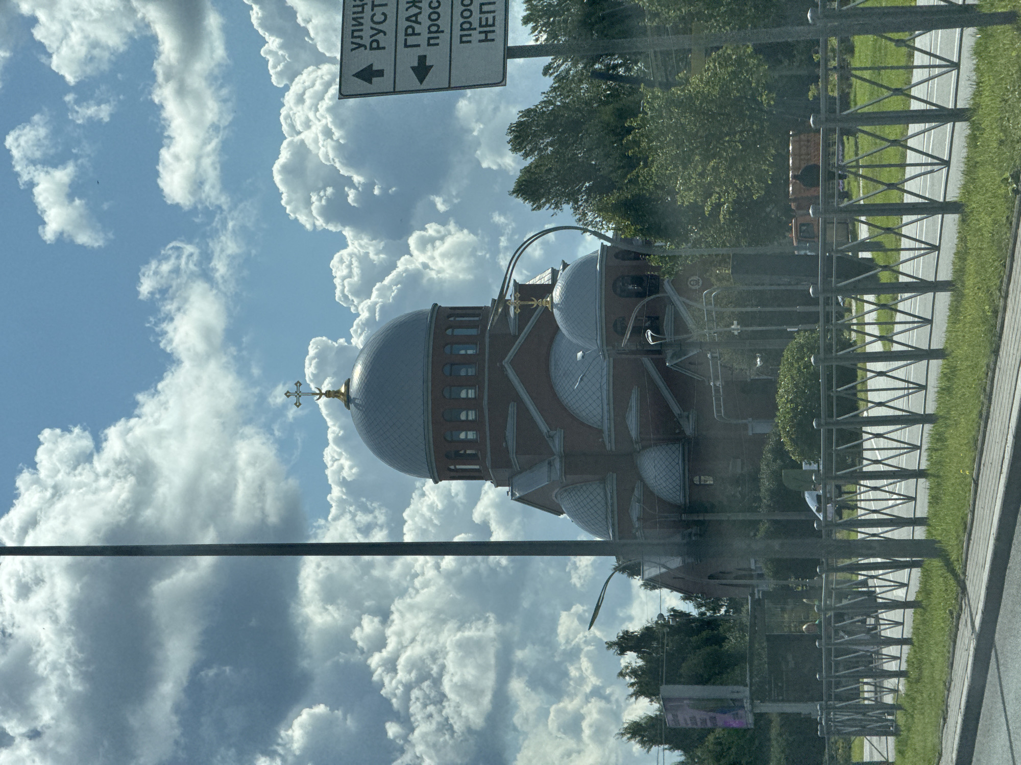 This image, taken in the city of Sinyavino, Russia, shows an Orthodox church with multiple domes, a tall central dome topped with a cross, and several smaller domes. The church is constructed from red brick and has a red tiled roof. In the foreground, a metal fence runs along a grassy area beside a road. A street sign with Cyrillic lettering is visible to the right of the church. The sky is partly cloudy with dramatic, fluffy white clouds against a blue sky. Trees and other vegetation are visible around the church and behind the fence. A tall, dark pole, possibly a lamppost or utility pole, is positioned in the left foreground. In the background, there appears to be a construction vehicle. The image appears to have been taken from a moving vehicle.