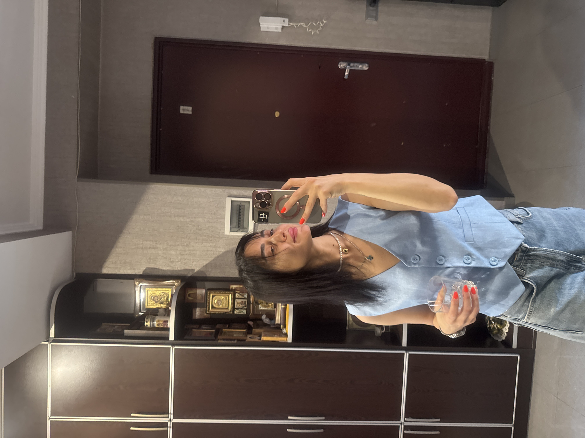 The image shows a woman taking a selfie in an indoor setting. The woman is wearing a light blue vest and denim jeans, and she has long dark hair. She is holding a clear glass in her right hand. Her nails are painted bright red. Around her neck, she is wearing a pearl necklace with a small cross pendant. In the background, there is a large, dark red door, a wall with textured wallpaper, and a dark brown cabinet with several shelves displaying framed items.
The scene appears to be a home or a private residence, possibly in Tbilisi, Georgia, given the location context. The lighting suggests it is daytime, but the exact time of day is not discernible. There are no immediate cues about the weather. The woman's expression seems relaxed and confident. The framed items on the shelves include what appear to be religious icons or artwork. On the phone screen, a Chinese character "中" is visible.