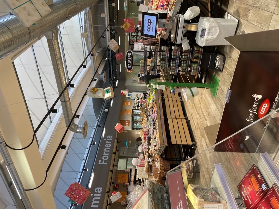 This media file captures the interior of a supermarket or grocery store in Castel del Piano, Italy, decorated with festive elements, suggesting a holiday season. The image is taken from a slightly elevated perspective, looking down an aisle or towards a main display area, and is rotated 90 degrees clockwise.
In the foreground, a large brown promotional display board is visible on the right, featuring the "fior fiore coop" logo and the text "Il Meglio della Cultura Gastronomica" (The Best of Gastronomic Culture). Below it, partially obscured, are packaged products, including "Salmone affumicato" (Smoked salmon). To the left, a long counter, likely part of a bakery or deli section, stretches into the background.
Overhead, a black track lighting system runs horizontally, from which multiple brightly wrapped gift boxes are suspended, adding a festive touch to the environment. Large, corrugated metallic air ducts are also visible along the ceiling. Below the lighting system, a prominent sign reads "Forneria" (Bakery).
Further into the store, various product displays are arranged on shelves. A digital screen displaying the "coop" logo and "Unione Amiatina" is positioned near a checkout or service counter area, alongside a digital scale and what appears to be a paper roll dispenser with a small bin. Another sign with "ITALPEPE" is visible multiple times on branding. A few blurry figures of shoppers or staff can be seen in the background near the counters and aisles. The flooring is a light-colored wood-like tile. The overall impression is a well-lit, organized retail space preparing for or engaging in a festive shopping period.
