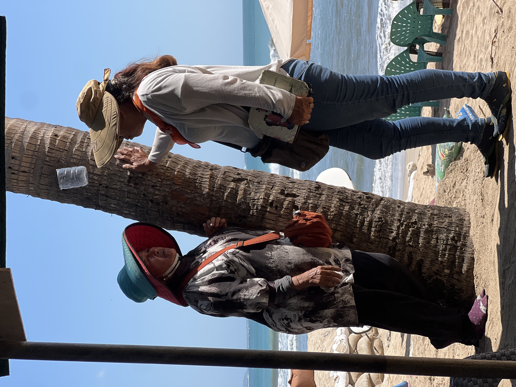 Two women are standing on a sandy beach in Kuta, Indonesia, by a palm tree. The woman on the left wears a wide-brimmed hat with a red interior and a dark, patterned long-sleeved shirt over black pants and patterned socks. She has an orange crossbody bag. The woman on the right wears a tan wide-brimmed hat, an orange scarf, a light-colored long-sleeved shirt, and blue jeans. She has a dark shoulder bag and holds a book. The ocean is visible in the background, with waves breaking on the shore. Several green plastic chairs are set up on the beach. The sky is clear and blue.