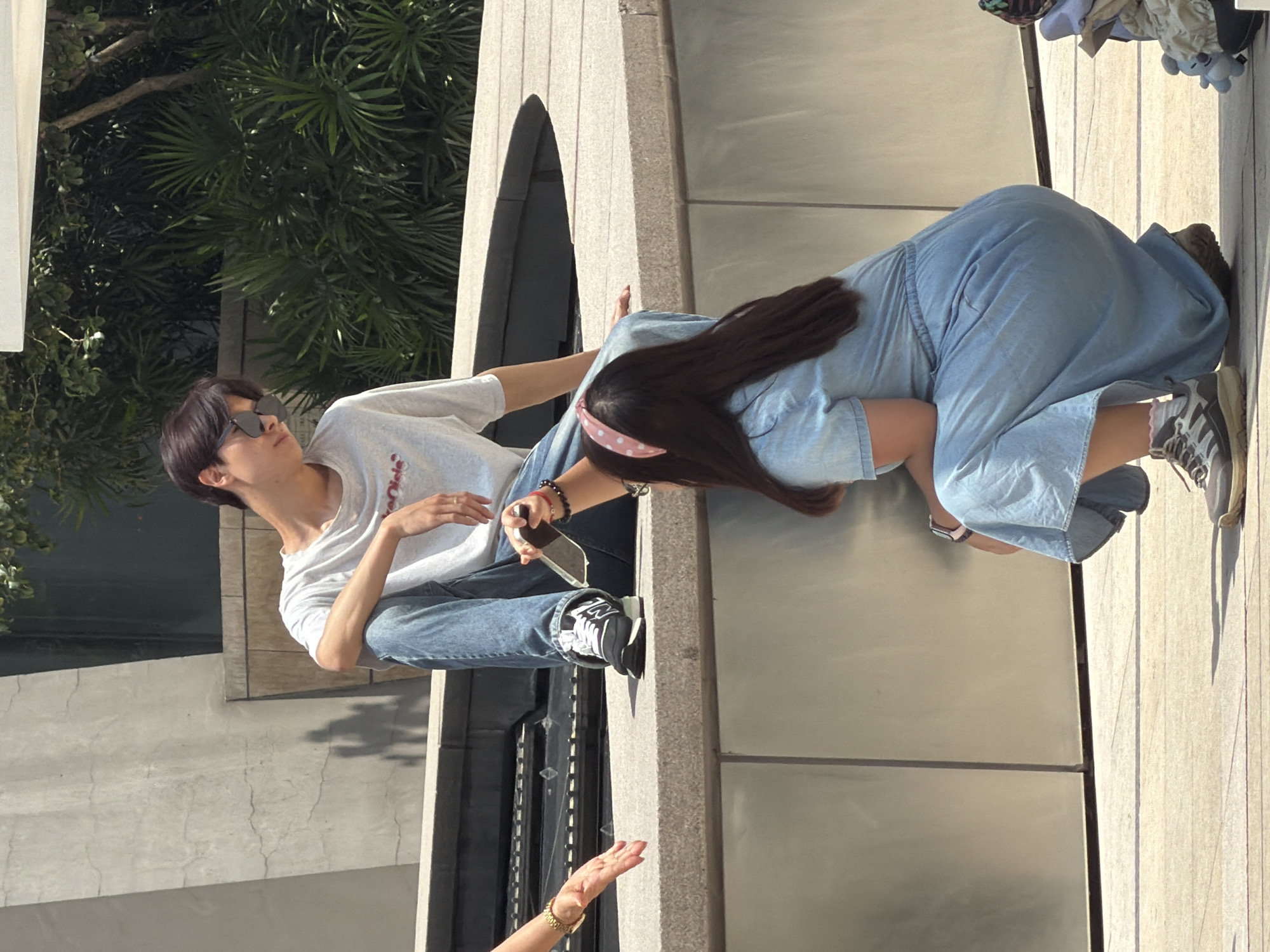 An adult male with dark hair, wearing sunglasses, a light-colored t-shirt, and blue jeans, is sitting on a ledge. He is looking down and to the right. An adult female with dark hair, wearing a light blue denim dress and a pink polka-dot headband, is crouching in front of him. She is holding a smartphone in her right hand and reaching towards him. Her left hand is resting on her leg. To the left of the male, another adult female's left hand with a gold bracelet is visible, reaching out. The environment appears to be an outdoor urban setting with concrete structures and lush green foliage in the background. The ground is paved with stone tiles.