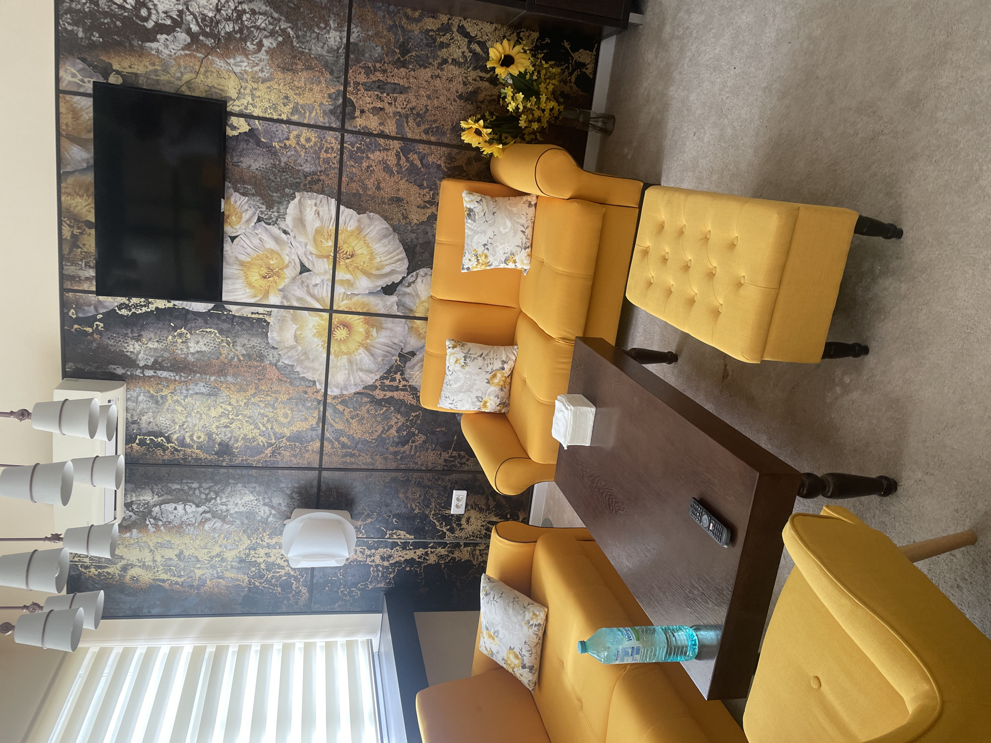 This media file captures an indoor scene, likely a living room or lounge area, characterized by a vibrant and modern aesthetic. The primary subjects are two plush yellow upholstered armchairs, each adorned with a decorative throw pillow featuring white, yellow, and gray floral patterns. A matching yellow upholstered ottoman or bench is positioned near one of the armchairs. A dark brown wooden rectangular coffee table sits in the center, displaying a stack of white tissues, a black television remote control, and a plastic water bottle with the partially visible label "V...l...a". Part of another yellow chair is visible in the foreground on the right.
The most striking feature of the room is a large wall art piece or wallpaper covering a significant portion of the back wall. It depicts a dark, textured background with shimmering gold abstract patterns, on which several large, luminous white and yellow poppy-like flowers are illustrated. A black flat-screen television is mounted on this decorative wall. In the upper right corner, a vase holds a cheerful bouquet of yellow sunflowers. The room is illuminated by a modern chandelier with multiple white conical shades and a smaller white wall sconce. The floor is covered with a light gray carpet, and windows are fitted with light-colored vertical blinds.
No people are present in the image, so no specific activity or event is taking place. The room appears prepared for relaxation or casual gathering. The dominant color palette of bright yellow and gold creates a cheerful and inviting atmosphere, balanced by the dark tones of the wall art and coffee table, lending a sense of warmth and modern sophistication. The lighting conditions suggest an indoor setting, but the exact time of day or outdoor weather cannot be determined.