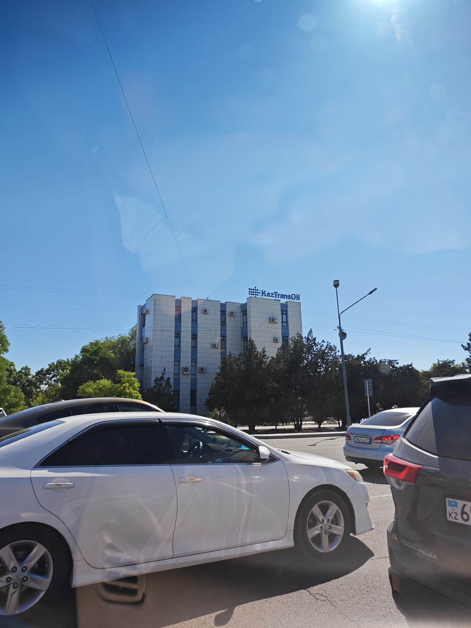The image shows a street scene in Aktau, Kazakhstan. In the foreground, a white Toyota Camry is parked or moving slowly. Behind it, the rear of a dark-colored SUV, possibly a Toyota Highlander, is visible. In the middle ground, a light-colored sedan with a license plate reading "C867ALD12" is seen. In the background, a modern, white and blue building with the "KazTransOil" logo is prominent. Several trees line the street, and electrical wires are strung across the sky. The sky is clear and blue, with the sun creating lens flares. The overall impression is a sunny day in an urban environment.
