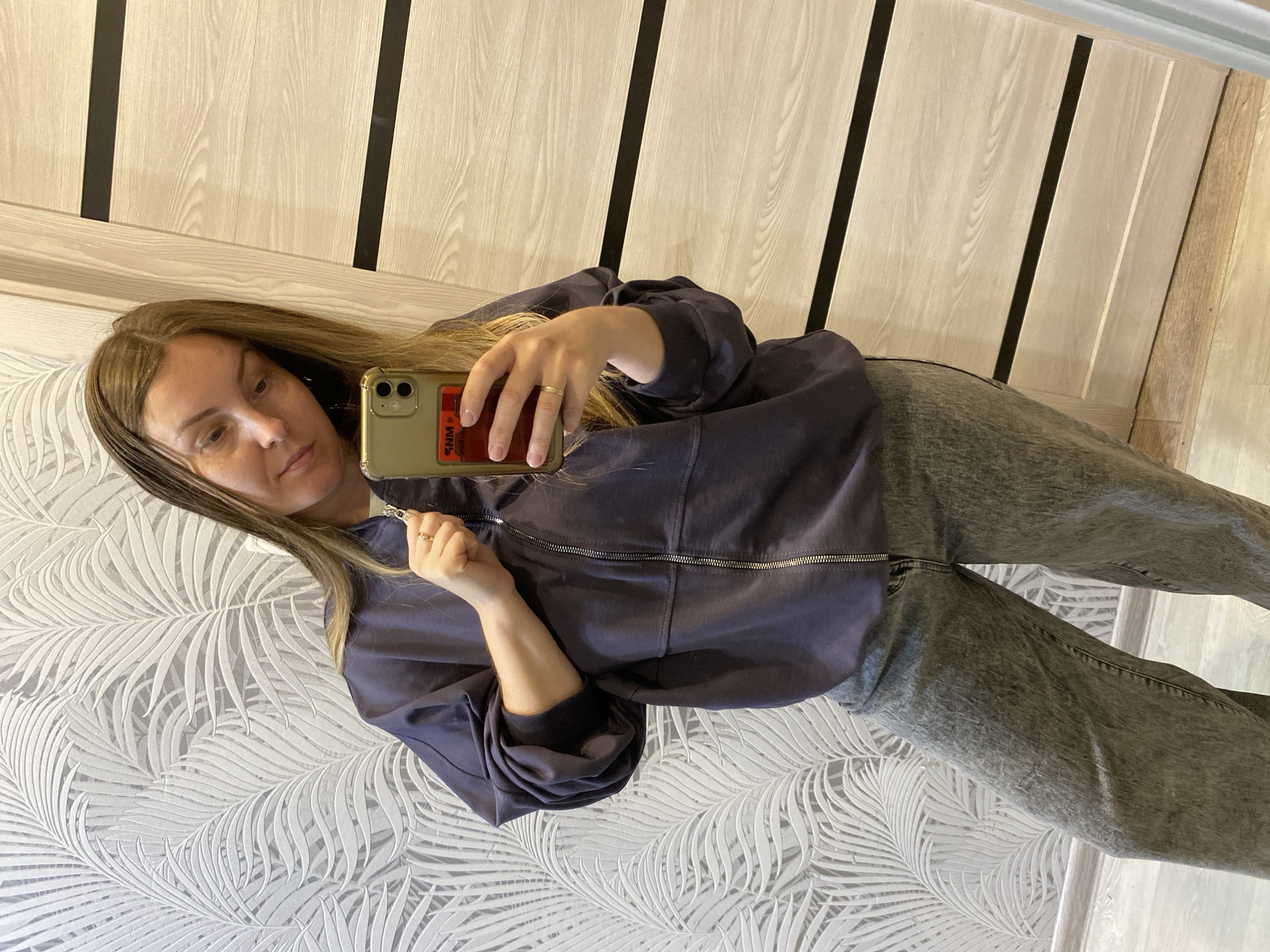 A woman with long brown hair is taking a selfie in front of a wall with a white and gray leaf pattern. She is wearing a dark blue zip-up jacket and gray jeans. She is holding a gold iPhone with a red card holder on the back. The background also includes a wall with vertical light brown and black stripes and a light brown floor. There is no information about the location being in Noviy, Russia.