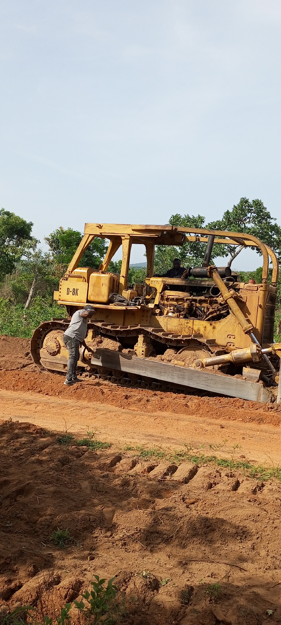 The image captures a daytime scene in what appears to be a rural or land-clearing area, likely Otunade, Nigeria, given the context. The central subject is a large, yellow track-type bulldozer, identified by the visible text "D-8K" on its side, and "CAT." on an interior part, indicating it's a Caterpillar D8K model.
Two men are present with the heavy machinery. One man, wearing a grey t-shirt, dark pants, and a dark cap, is standing beside the bulldozer's tracks, appearing to inspect or work on the lower part of the machine. The other man, dressed in a dark shirt, is seated in the operator's cabin of the bulldozer, suggesting he is either preparing to operate it or is currently operating it.
The bulldozer is situated on reddish-brown, disturbed earth, indicative of recent ground work or a construction site. Clear track marks are visible in the soil around the machine. In the background, there is abundant green foliage and trees under a bright, clear, light blue sky, typical of good weather conditions. The scene implies ongoing land development or maintenance activities. No strong emotions are discernible from the individuals.