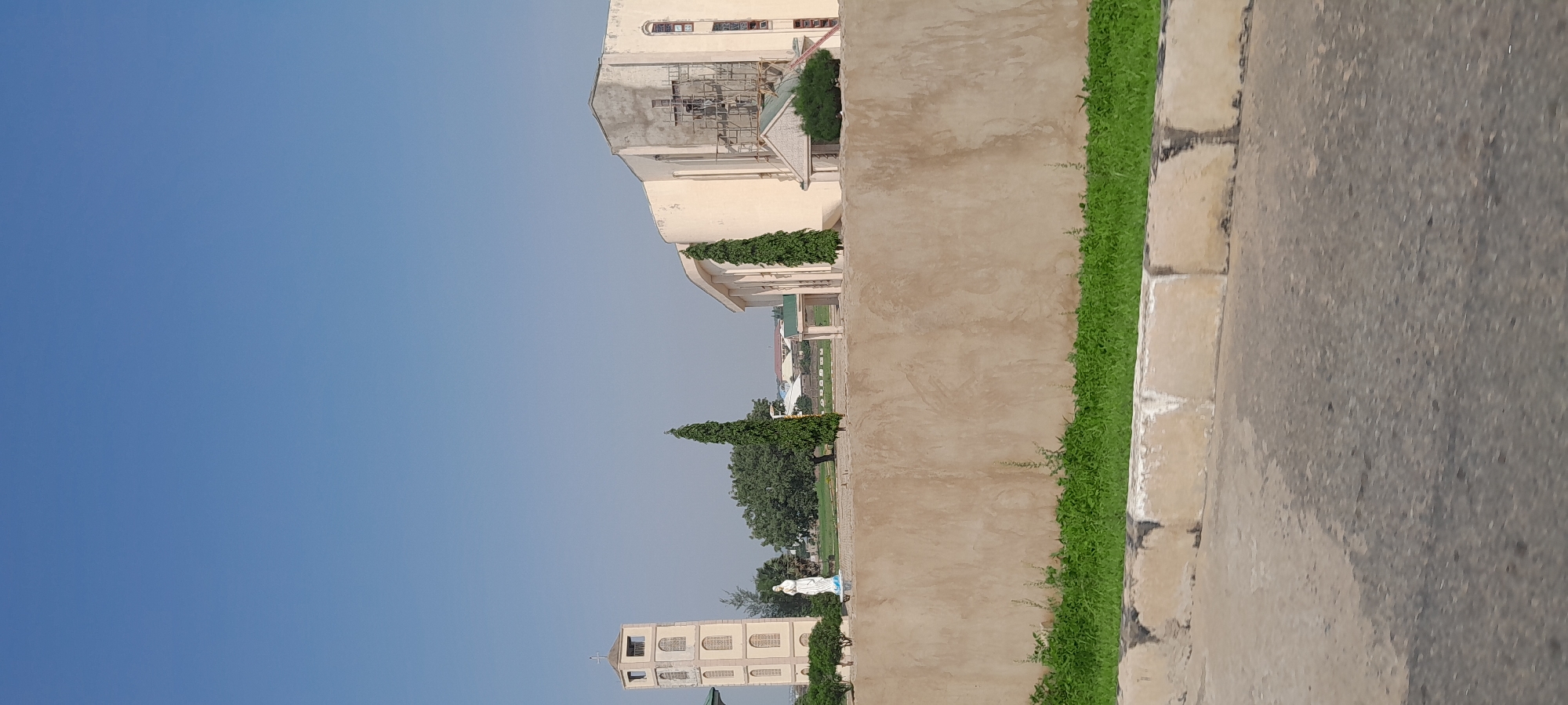 The image captures a daytime exterior view of a church complex, likely located in Jalingo, Nigeria. A prominent bell tower stands to the left, constructed of light-colored stone with arched openings. In front of the tower, a white statue of a person, possibly Mary, is visible. Adjacent to the tower, a large building, presumably the main church structure, features a curved facade and appears to be undergoing some construction or renovation, with scaffolding visible on one side. Two tall, slender green trees stand in front of this building. A long, light brown wall dominates the middle ground, separating the street from the church property. Green grass grows along the base of this wall. The foreground shows a paved road with a concrete divider. The sky is clear and blue.