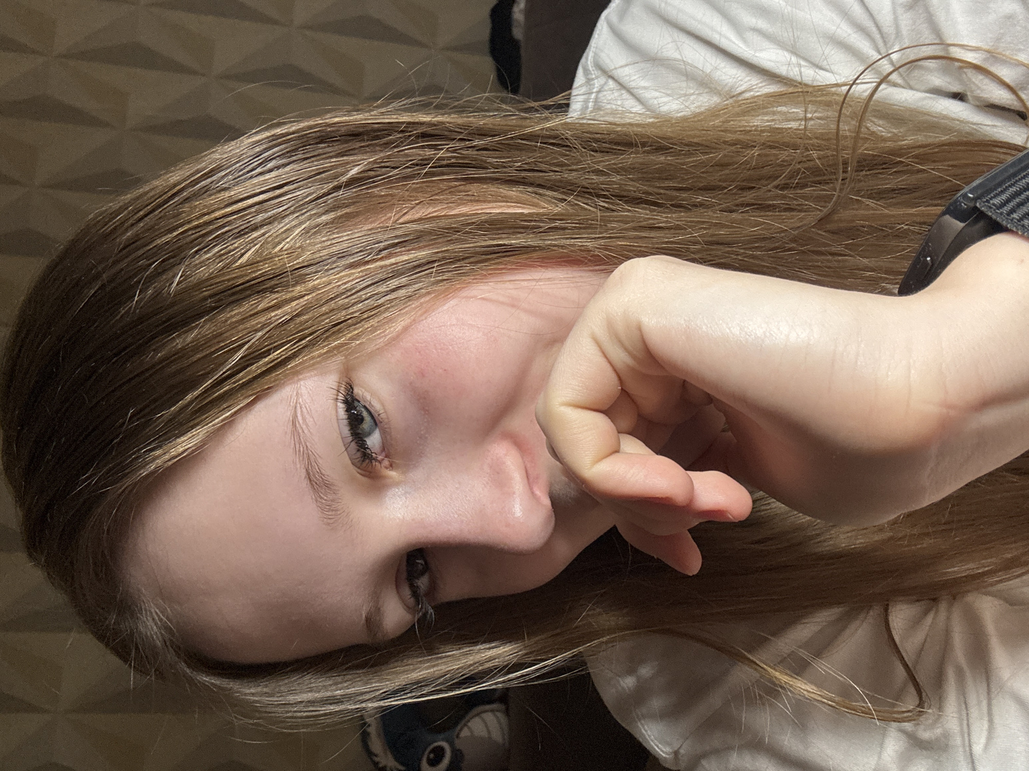 A close-up portrait of a light-skinned woman with long, straight blonde hair. She is positioned at an angle, with her head tilted to the left. Her left eye is more prominent in the frame, revealing a light blue iris. She is touching her lips with her fingers. Her hair is slightly oily in appearance. A black watch is visible on her wrist. She is wearing a white shirt. The background shows a textured, geometric pattern, possibly a wall. An object with eyes is on the left part of the image. There is no indication of the city of Chaikovsky, Russia, in the image.