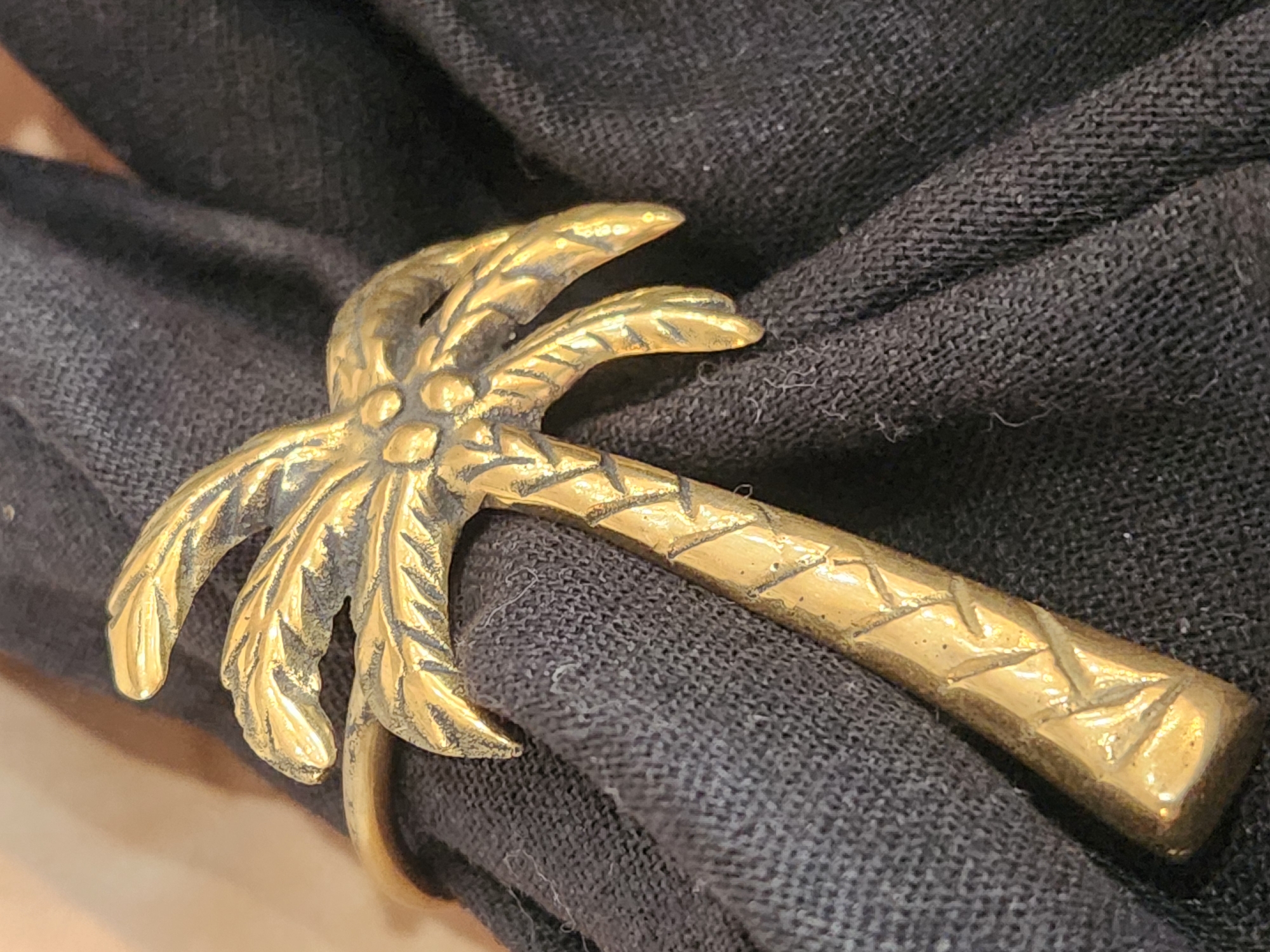 A close-up, eye-level shot focuses on a gold-colored ring shaped like a palm tree. The ring is worn on dark grey fabric, which appears to be a napkin or cloth, bunched and folded. The palm tree design is detailed, with individual fronds and a textured trunk. The lighting creates highlights and shadows on the metallic surface, emphasizing the intricate design. The background is out of focus, showing a muted, warm tone, possibly a wall or countertop. There are no people or animals visible in the image. The scene is indoors, likely in a domestic setting, but no specific location in Maitland, United States of America can be determined from the visual information.
