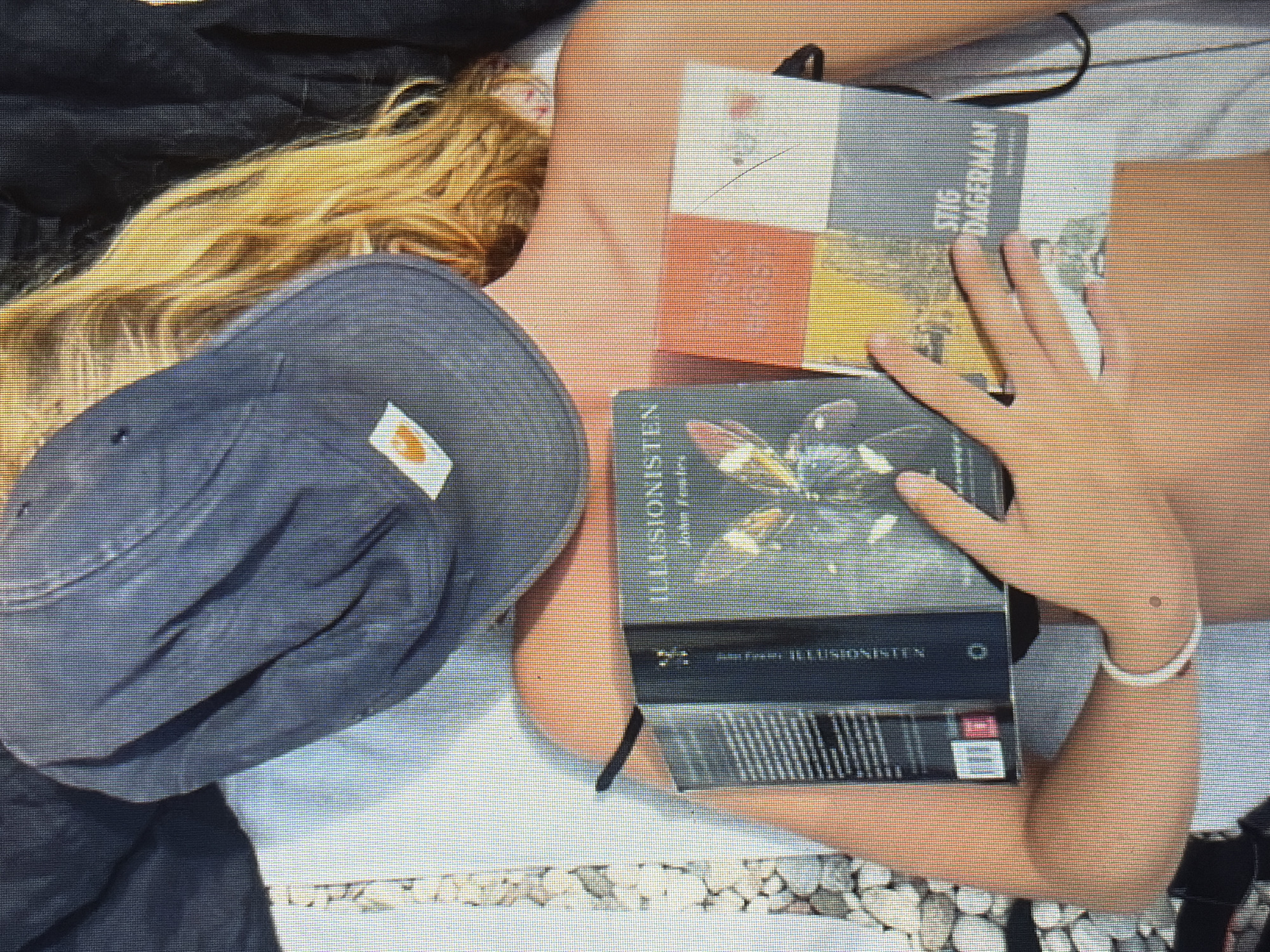 A person is lying down, wearing a blue baseball cap that covers their face. They have blonde hair and are wearing a white bracelet on their left wrist. They are holding three books. The book on the left is black with text and an image on the cover. The middle book is dark with an illustration of a creature on the cover. The book on the right has a cover divided into red, yellow, and blue sections, with text and an image. The person is lying on a white surface, possibly a bed or a blanket, with some stones visible on the left side of the image. The background is dark. The scene is outdoors, likely in a park or garden, given the presence of stones and vegetation.