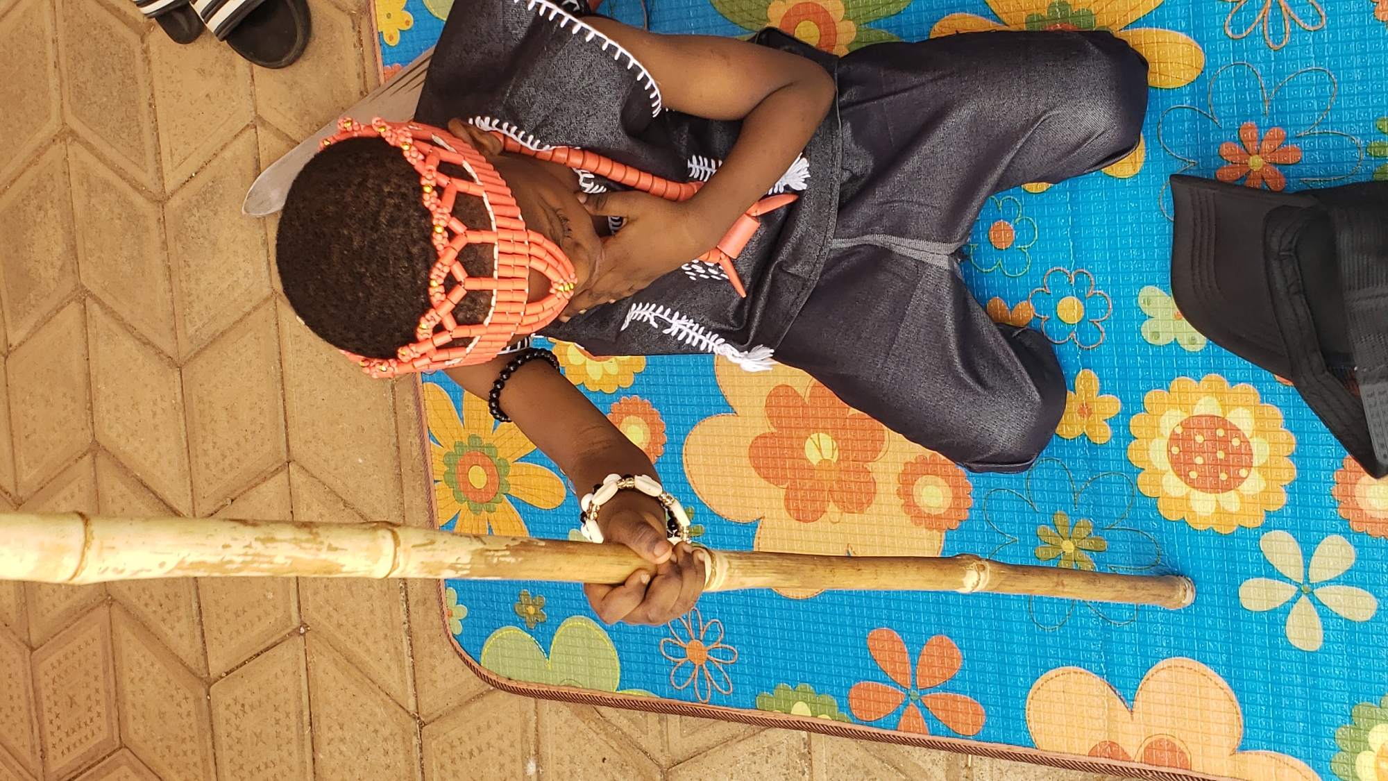 A person wearing traditional clothing, including a coral beaded headpiece and necklace, lies on a blue and orange floral patterned mat. The person is holding a light brown wooden staff. The location appears to be in Sabon Tasha, Nigeria, based on context. The person also wears black and white beaded bracelets. Part of a pair of black and white striped sandals are visible at the top of the frame. A black fabric item rests on the mat to the right. The floor visible beneath the mat is composed of beige-colored interlocking pavers.
