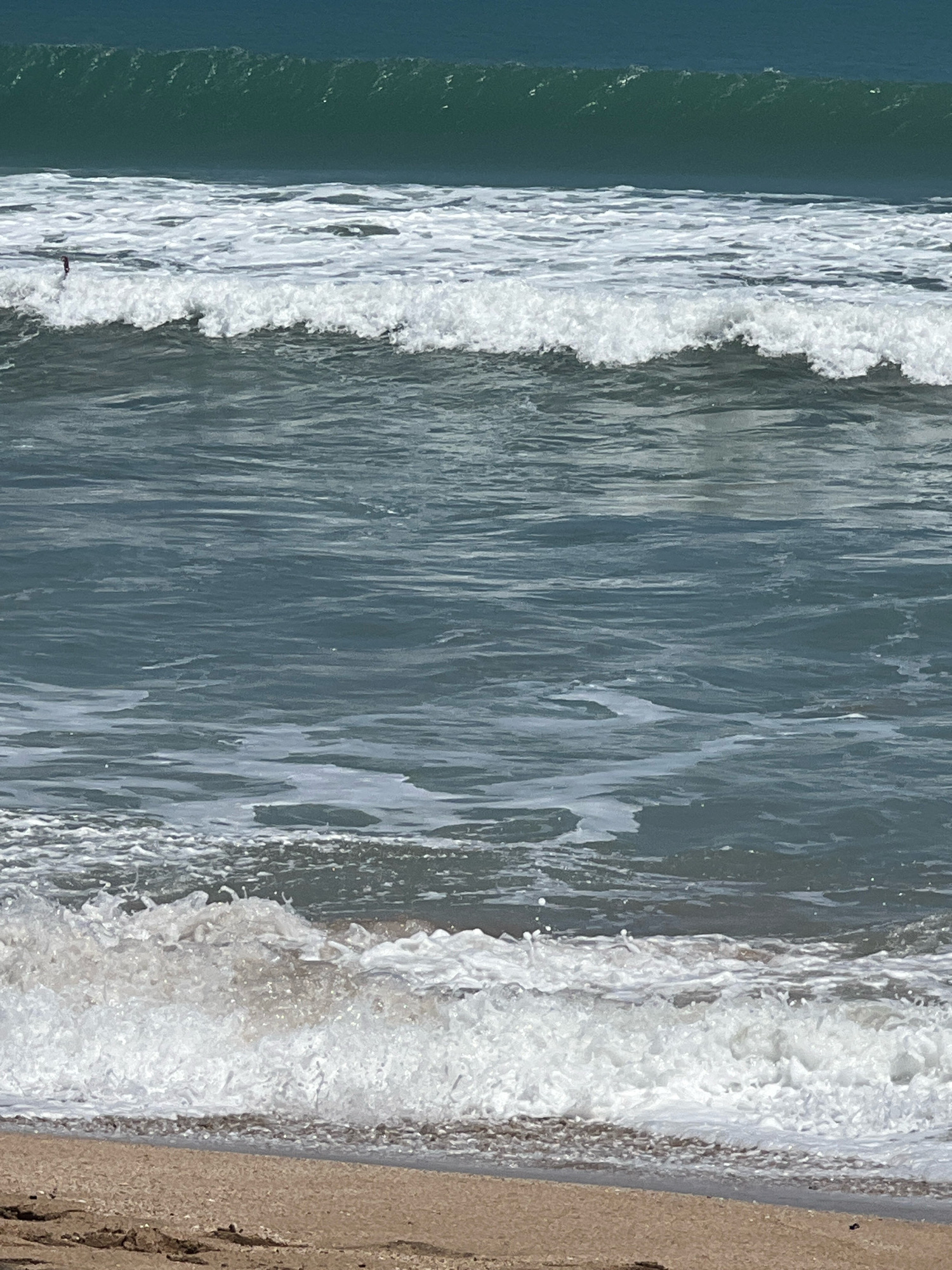 The image captures a view of the ocean waves breaking on a sandy beach. The water is a deep teal color in the distance, transitioning to lighter blues and whites as it approaches the shore. Several waves are visible, with the closest one actively cresting and breaking into foam. A small, dark object, possibly a person on a surfboard, is discernible in the distance within a wave. The foreground shows a sandy beach with some debris scattered. The scene depicts a typical oceanfront environment, likely a popular surfing or beach destination. Without additional information, the specific location in Kuta, Indonesia, cannot be definitively identified solely from the visual elements.