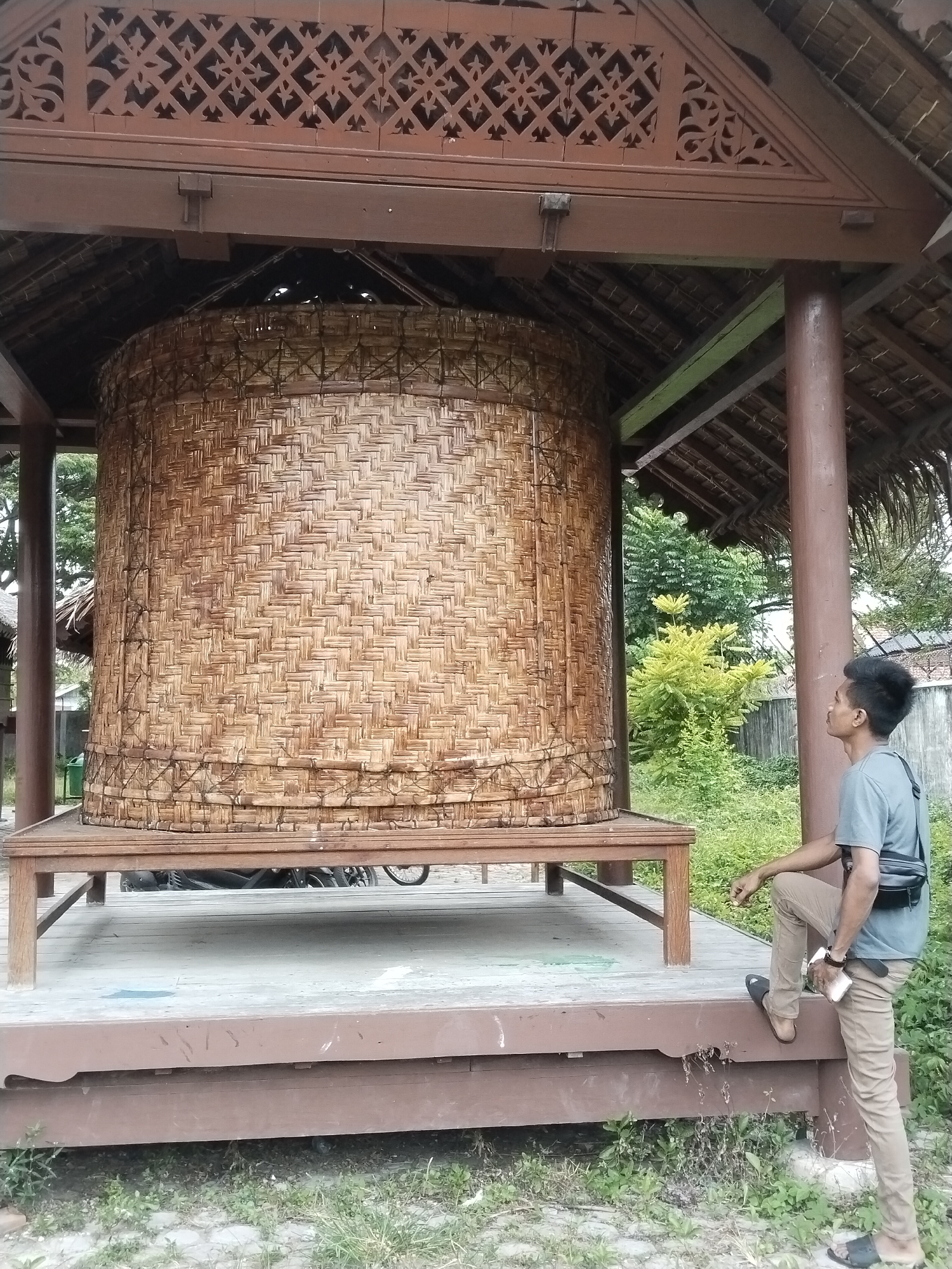 The image shows a large, cylindrical, woven structure housed within a wooden pavilion. This structure is identified as a traditional rice storage container. A man is standing on the pavilion's edge, looking up at the woven container. He is wearing a grey t-shirt, khaki pants, and sandals, and has a black shoulder bag. The pavilion features decorative wooden carvings along the upper edge. The location is Lhokseumawe, Indonesia. Vegetation, including trees and shrubs, is visible in the background, partially obscured by a fence. A bicycle is underneath the pavilion. The day appears overcast.