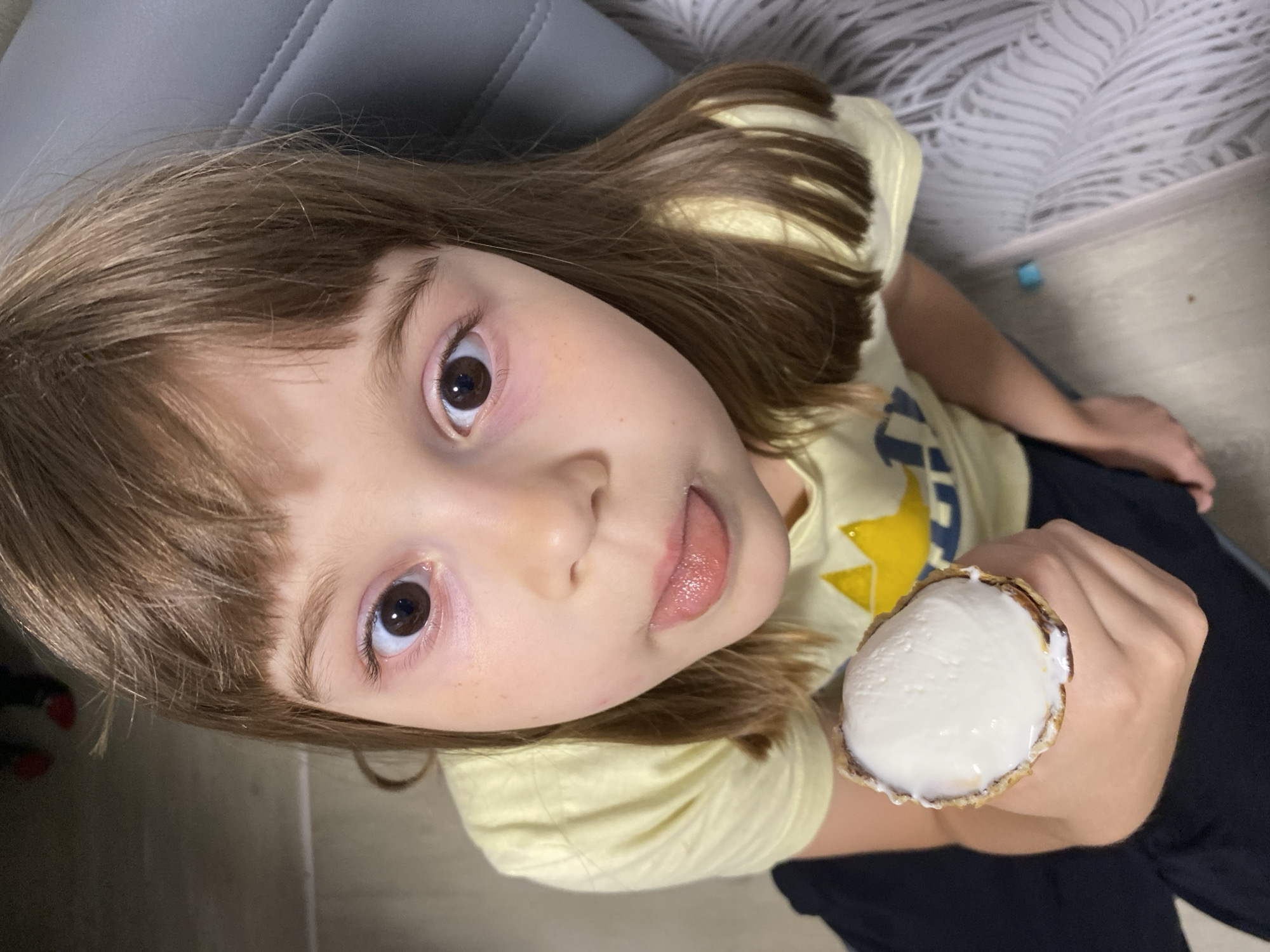 The scene depicts a child indoors in Noviy, Russia. A young individual with light brown hair and bangs is positioned centrally, looking directly at the camera with wide, open eyes and their tongue slightly extended. The child wears a light yellow short-sleeved t-shirt featuring a prominent yellow and blue star design on the front, and dark-colored trousers. In the child's right hand, an ice cream cone is held, containing a scoop of white ice cream. The waffle cone exhibits dark internal coloration, suggestive of chocolate lining or melted ice cream residue. The background to the immediate left of the child consists of a dark, uniform surface, possibly a floor or wall. In the upper left quadrant, red footwear is partially visible on a dark ground. To the right of the child, a grey upholstered surface with visible stitching, indicative of furniture such as a couch or chair, is present. Below this furniture, a light-colored textile with a wave-like pattern is visible on the floor, alongside a small blue object in the lower right corner of the frame.