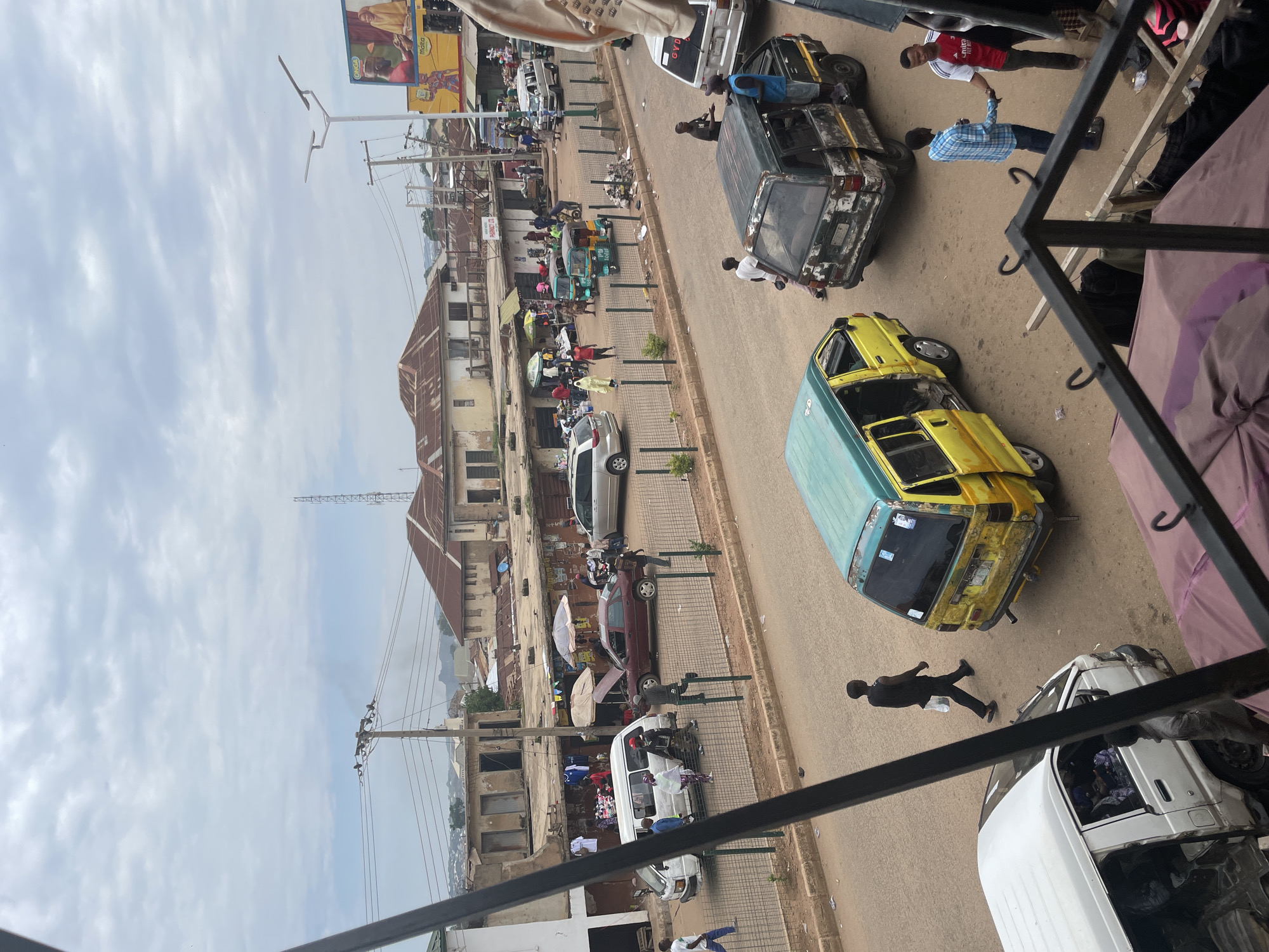This image depicts an overhead view of a bustling street scene in the city of Unknown, Unknown, likely a market or commercial area, under an overcast sky. The road surface appears unpaved or very worn, with visible dust and dirt.
In the background, a cluster of single and two-story buildings with aged, reddish-brown corrugated metal roofs lines the street. Many structures show signs of wear and disrepair. A tall communication tower is visible on the left horizon, and distant hills are discernible. Numerous power lines crisscross the upper portion of the frame, connected to wooden utility poles. On the far right, a large yellow billboard displaying "ONGA" and "Malta" logos with images of people stands next to a modern street light featuring two horizontal lamp arms.
The midground features a two-lane road divided by a low metal barrier fence. On the left side of the fence, a white van with its driver's door open is parked, with several people standing around it, one appearing to open the rear door. Further right along the fence, a maroon sedan is stopped with its hood open, suggesting a breakdown or maintenance, surrounded by a few individuals. Adjacent to the fence and extending into the background are several makeshift market stalls with fabric umbrellas, where goods, including clothing, are displayed. Further down the street, a silver SUV is parked, and at least two three-wheeled motorized rickshaws (keke napep) are visible. People are walking and standing throughout this area, engaging in various activities such typical of a market environment.
In the foreground, closer to the viewer, the scene is dominated by several commercial vans. On the far left, a white van is partially visible. Centrally, a worn yellow and teal van with its passenger doors open is positioned on the road. To its right, an even more heavily used dark green van, possibly modified, also has its doors open, and a person in a blue shirt is standing on its side or step. A man in a black shirt and pants walks across the street from left to right. On the far right, two men are shaking hands; one wears a blue plaid shirt, and the other a red and white football jersey with "Emirates FLY BETTER" on it. Part of a dark metal railing and a folded purple/brown tarp, indicating the elevated vantage point of the photographer, occupy the bottom edge of the frame.