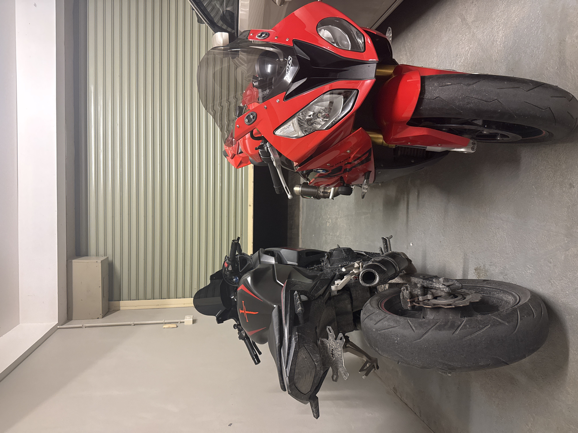 Two sport motorcycles are parked side-by-side indoors, in front of a closed metal roll-up garage door. The motorcycle on the left is black with red accents, featuring a stylized red cross on the fuel tank and a custom license plate bracket. It is viewed from the rear, showcasing its large rear tire and exhaust. The motorcycle on the right is bright red with black accents, its front end prominently displayed. It has a tinted windscreen and gold-colored front forks. A beige wall is visible to the left, and a hint of a light-colored car is present on the far right. The floor appears to be concrete.