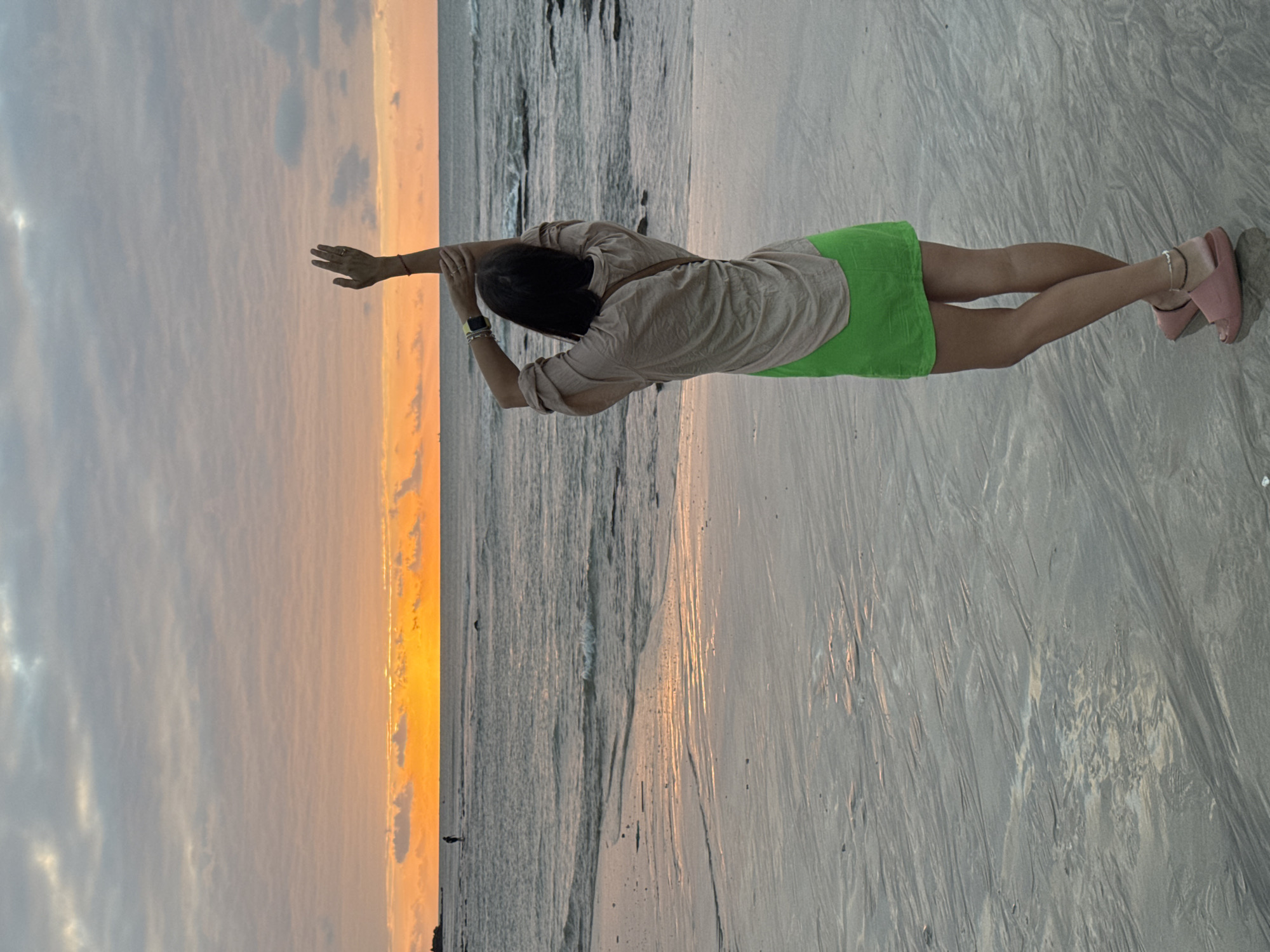 A lone person stands on a wet sandy beach facing the ocean at sunset. The person is wearing a light-colored loose-fitting shirt and a bright green skirt. Their arms are raised above their head. The sky is filled with clouds reflecting the orange and yellow hues of the setting sun. The ocean is calm with gentle waves lapping at the shore. The wet sand shows patterns from receding water. In the distance, a solitary figure walks along the water's edge. The location is Kuta Selatan, Indonesia.