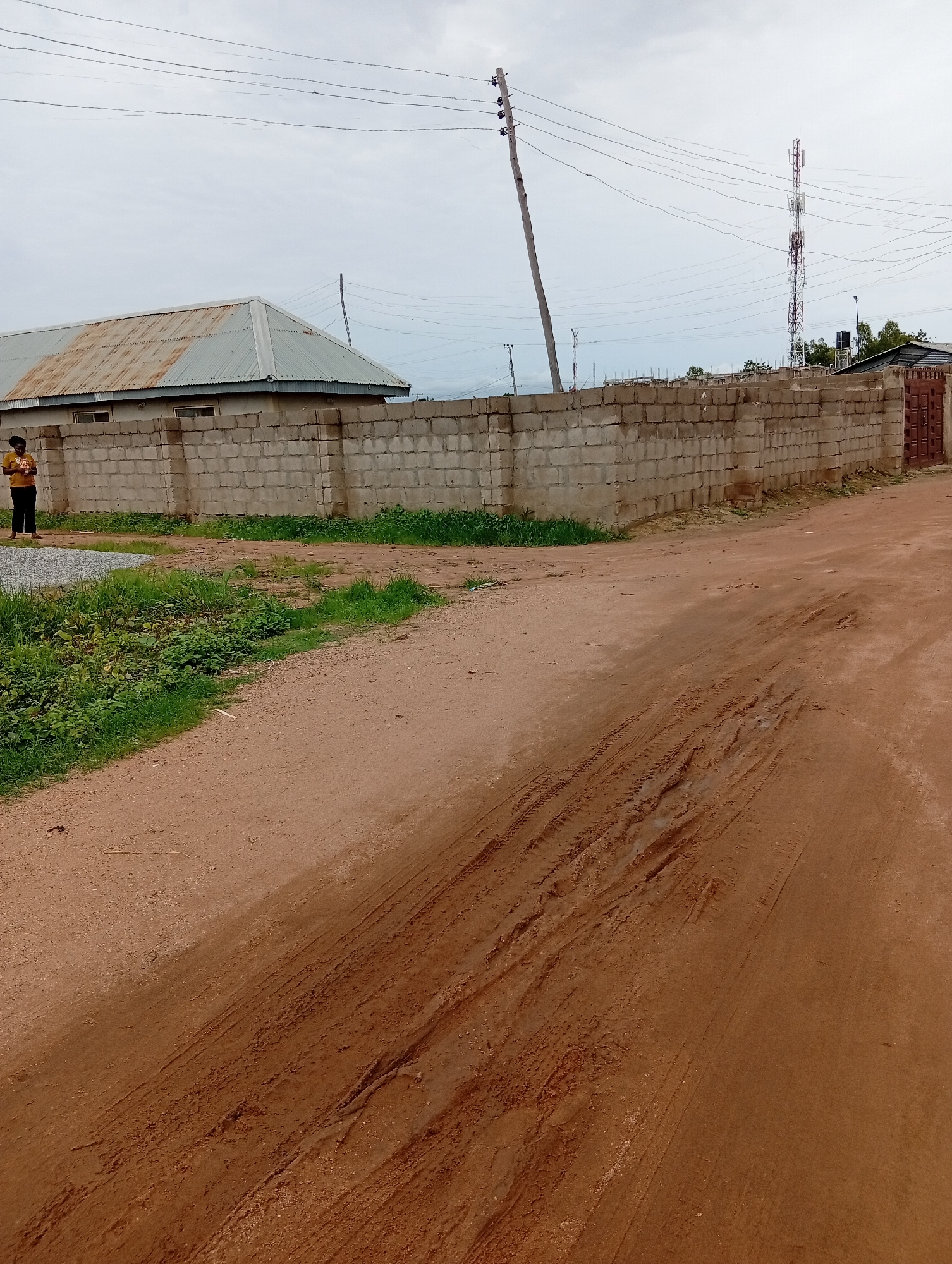 The image depicts an outdoor scene in Jalingo, Nigeria, characterized by a dirt road with prominent tire tracks, indicative of recent vehicular activity. To the left, a low building with a corrugated metal roof and concrete block walls is visible, with a person standing near its entrance, wearing an orange t-shirt and dark trousers. A low concrete block wall runs along the front of the building and extends to the right, forming a corner. Beyond this wall, a tall telecommunications tower is visible in the distance, a common feature in urban and semi-urban landscapes. The environment appears to be a developing area, with construction materials and overgrown vegetation present. The sky is overcast, suggesting a cloudy day.