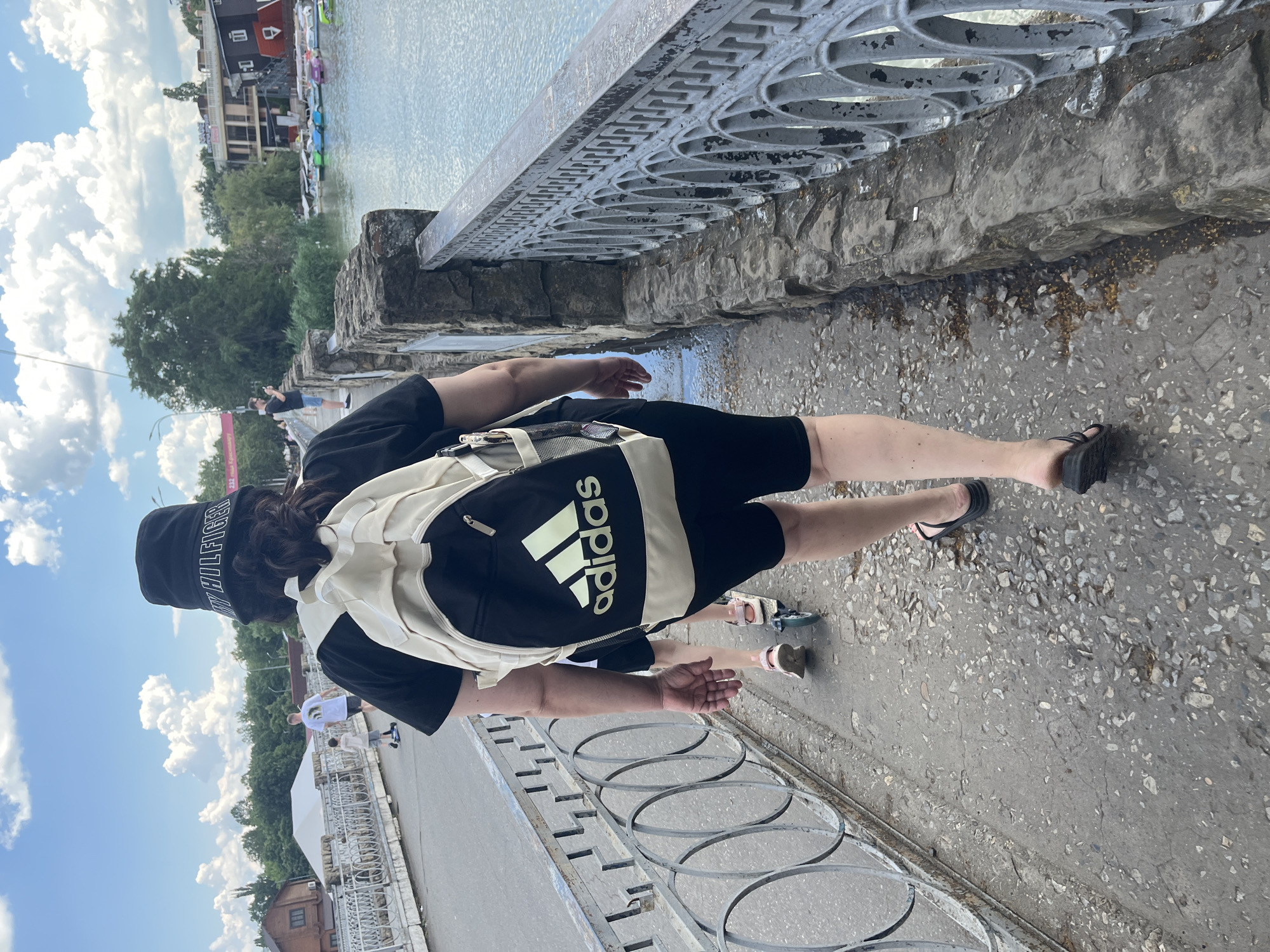 The image captures a scene on a bridge in Krasnodar, Russia, on a partly cloudy day. In the foreground, a person is walking away from the camera, wearing a black bucket hat, a black t-shirt, black shorts, and black flip-flops. They are carrying a beige and black Adidas backpack. To the left of this person, another individual, possibly a child, is riding a scooter. The bridge has a weathered concrete surface with scattered debris and a metal railing. On the right side of the bridge, a stone wall lines the waterway. Across the water, buildings and trees are visible, and a variety of colorful pedal boats are moored. The sky is a mix of blue and white clouds, indicating fair weather.