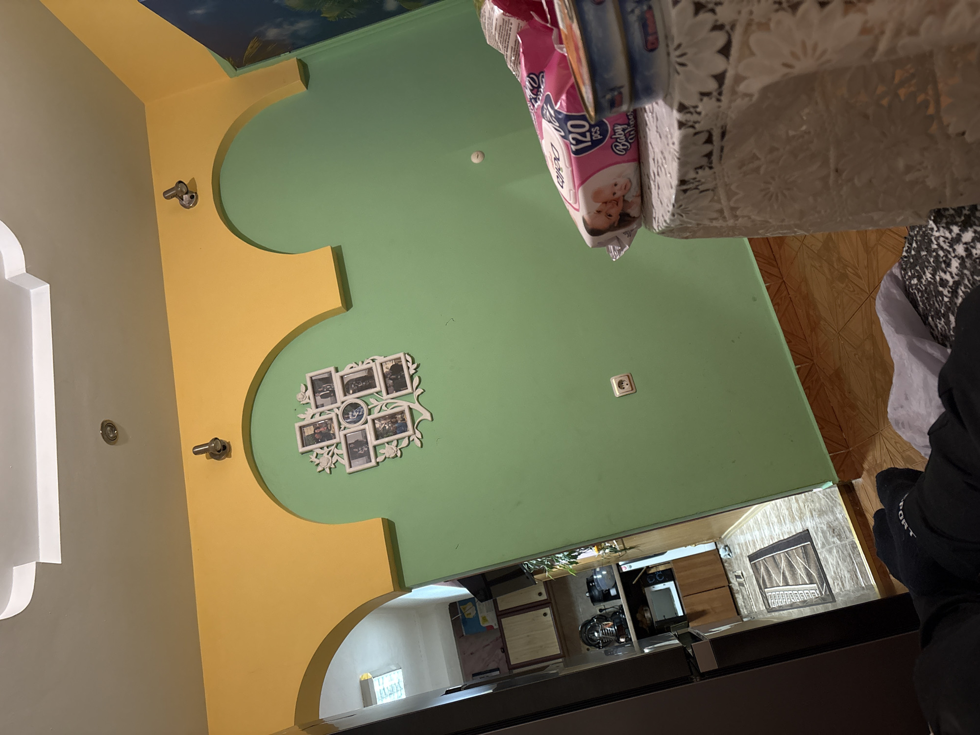 The image captures an interior residential space, likely a living or dining room, connected to a kitchen area. The room features a distinctive wall design with segments of light beige/grey, bright yellow arched trim, and light green paint. A partial blue sky mural adorns a section of the ceiling in the upper left.
Decorative elements include two metallic hooks or fixtures on the yellow trim, and a white, vine-shaped picture frame displaying multiple small photographs on the green wall. A chrome fixture is also present on the beige/grey wall, and a single European-style electrical outlet is visible on the green wall.
In the immediate foreground, a table draped with a white, lacy tablecloth holds a pink package of "Baby Wipes" marked "120 PCS" and featuring an image of two infants. Another object, possibly a book, rests beside the wipes. A hand, clad in a black glove with the partial word "PORT" visible, is seen in the bottom right, resting on a dark surface.
Beyond an archway on the right, a kitchen or utility area is visible, equipped with white appliances, wooden cabinetry, and a patterned, wood-effect tiled floor. A further archway in the background leads to another brightly lit room with a window. The scene suggests a lived-in home environment in Dolna Banya, Bulgaria.