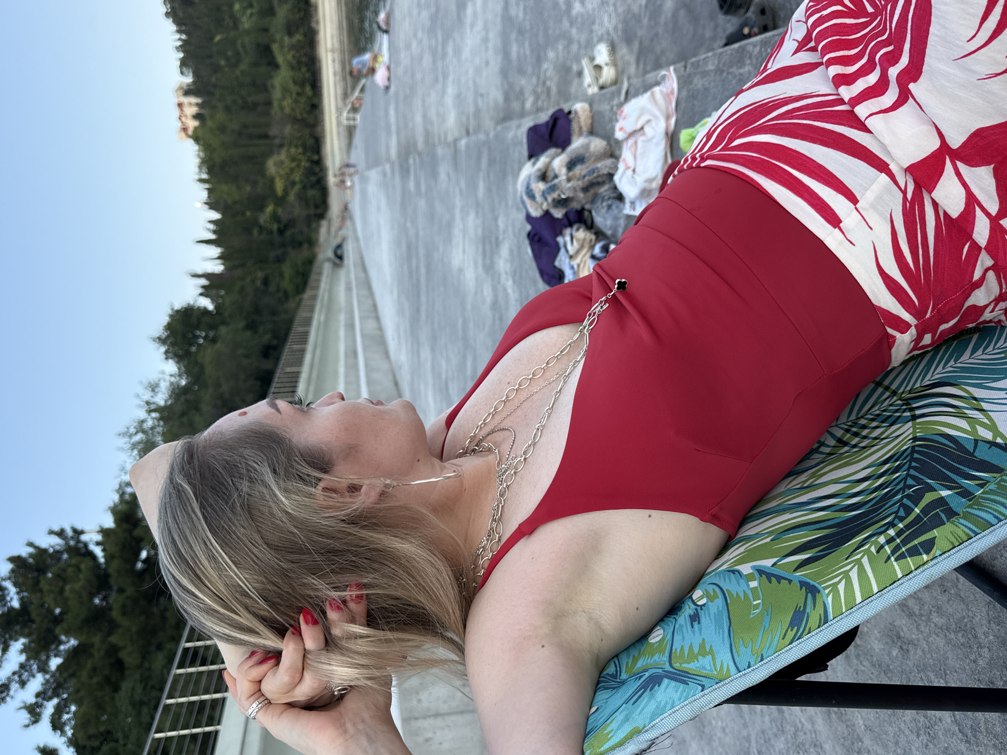 A woman with blonde hair is seated on a colorful beach chair, looking to her right. She is wearing a red one-piece swimsuit and a white skirt with red palm leaf patterns. Her right arm is raised, with her hand resting on the back of her head, and she is adorned with several silver necklaces and large hoop earrings. The scene appears to be a public walkway or pier made of concrete, situated by a body of water, possibly the sea. In the background, there are trees and a building on a hillside, suggesting a coastal resort area. Other people are visible in the distance, some appearing to be relaxing or swimming. The overall atmosphere is casual and relaxed, indicative of a leisurely day outdoors. The image does not provide specific details to confirm the location as Utios, Ukraine.