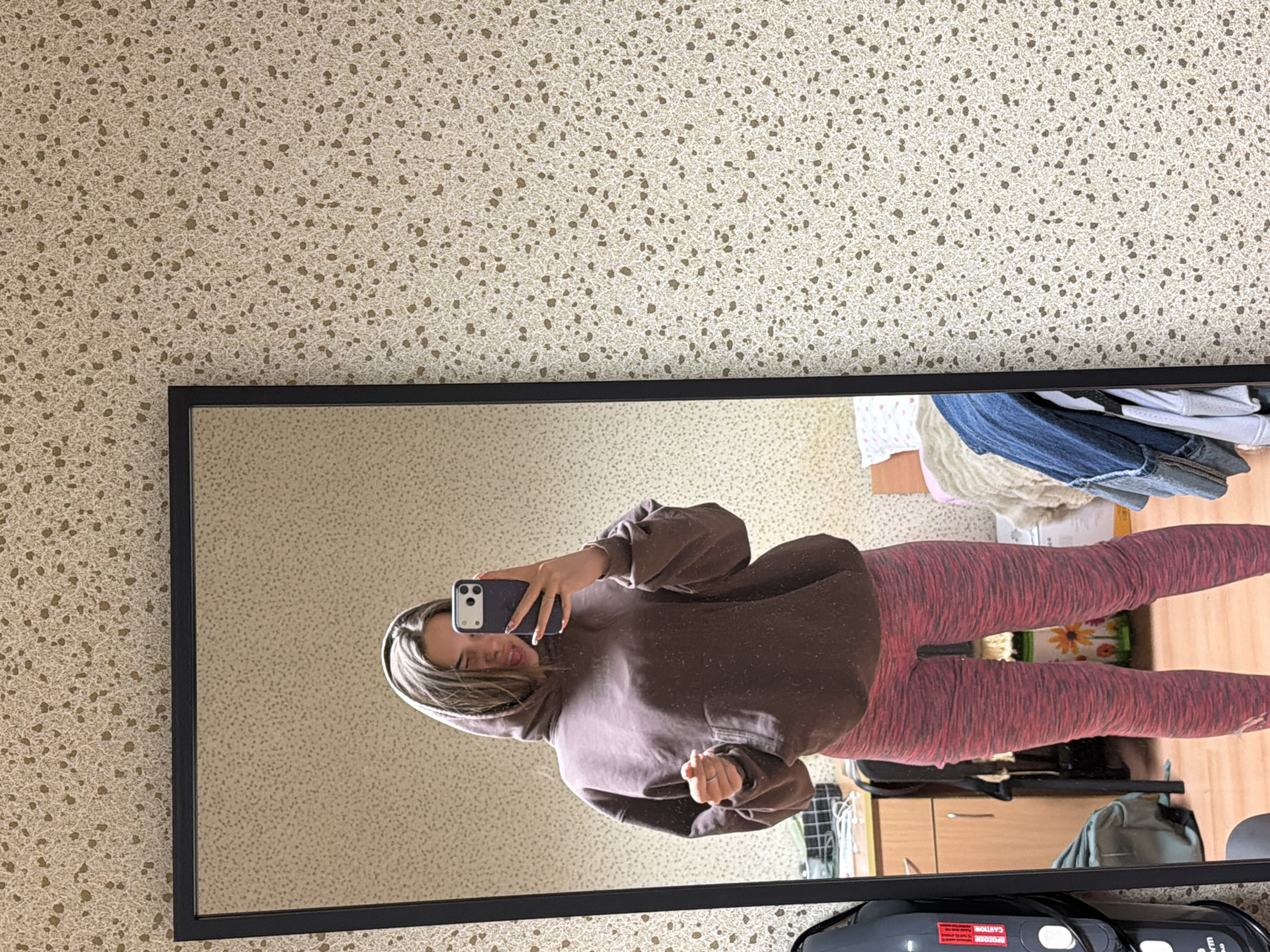 A young woman is captured in a mirror selfie indoors, appearing to be in Sofia, Bulgaria. She stands facing the mirror, holding a dark-colored smartphone, likely an iPhone with dual cameras, to take the picture. Her right hand is visible holding the phone, while her left hand is partially seen clenched near her stomach. She is wearing an oversized dark brown hooded sweatshirt and form-fitting red or pink patterned leggings. Her hair is long, light brown or blonde with highlights, framing her face. She has a subtle smile or smirk, and her eyes appear squinted or closed playfully.
The mirror has a thin black frame and reflects a room with light-colored walls covered in a speckled wallpaper pattern of beige or off-white with irregular brown dots. In the background reflected behind the woman, a cluttered domestic space is visible. To her right, a pile of clothes, including blue denim jeans, rests on top of what appears to be a wooden dresser or cabinet. To her left, part of a white bag or laundry basket is visible. In the foreground, at the bottom of the mirror's reflection, a black appliance, possibly a humidifier, is present. Text on this object includes "CAUTION" and partially visible words like "TOXICUM" and "warm". The overall impression is that of a casual moment captured in a lived-in room.