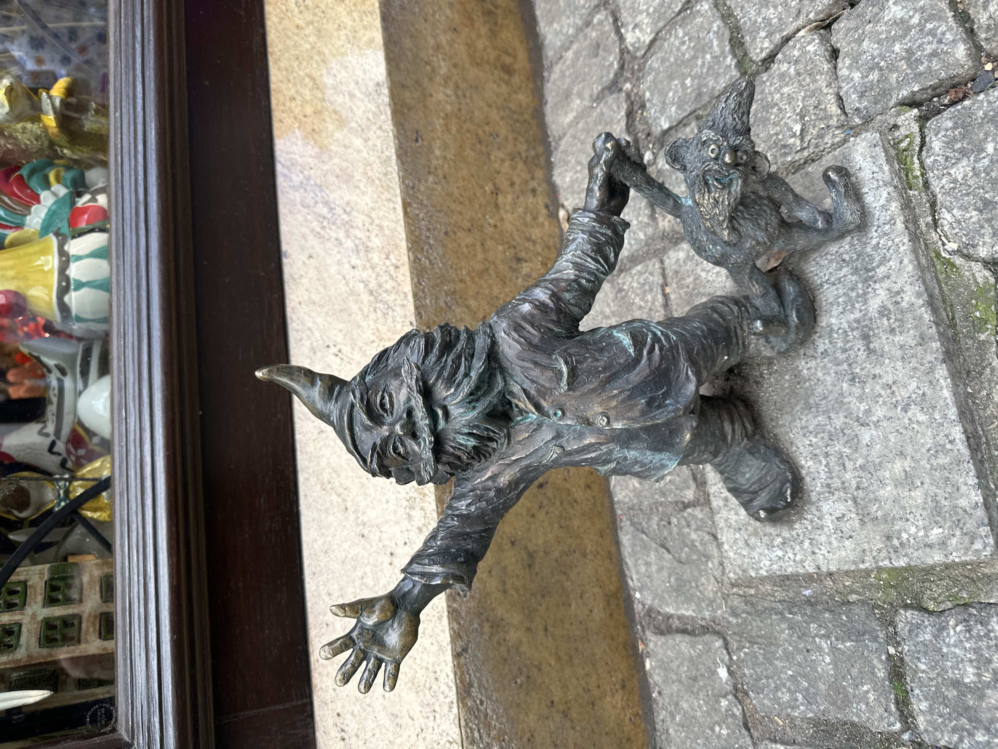 The image shows two small bronze gnome statues in Wrocław, Poland. One gnome, positioned on the ground, extends its arm towards another gnome situated on a beige, vertical surface, possibly a step or wall. They appear to be holding hands. The ground is paved with grey stone. The edge of a shop window with miscellaneous items inside is visible to the left.
