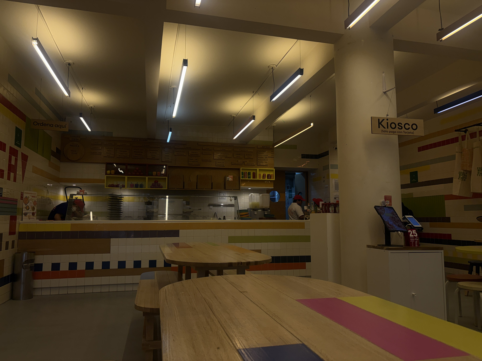 This is an interior shot of a restaurant or eatery, possibly located in Mexico City, Mexico, based on the Spanish signage. The space is well-lit with several fluorescent light fixtures suspended from the ceiling. The focus is on the seating area, with wooden tables and benches dominating the foreground. The tables are decorated with colorful stripes.
In the background, there is a service counter where food preparation appears to be taking place. Two individuals, one in a red hat, are visible behind the counter. The walls are decorated with colorful tiles and signage in Spanish.
There are tables and chairs to the right of the main pillar. A sign above the pillar reads "Kiosco (Sólo pago con Tarjeta)", which indicates that only card payments are accepted. Next to this is a self-ordering kiosk with a screen, indicating the digital nature of the place. A trash can stands in the corner, and the flooring appears to be light grey. The overall environment suggests a casual dining space.