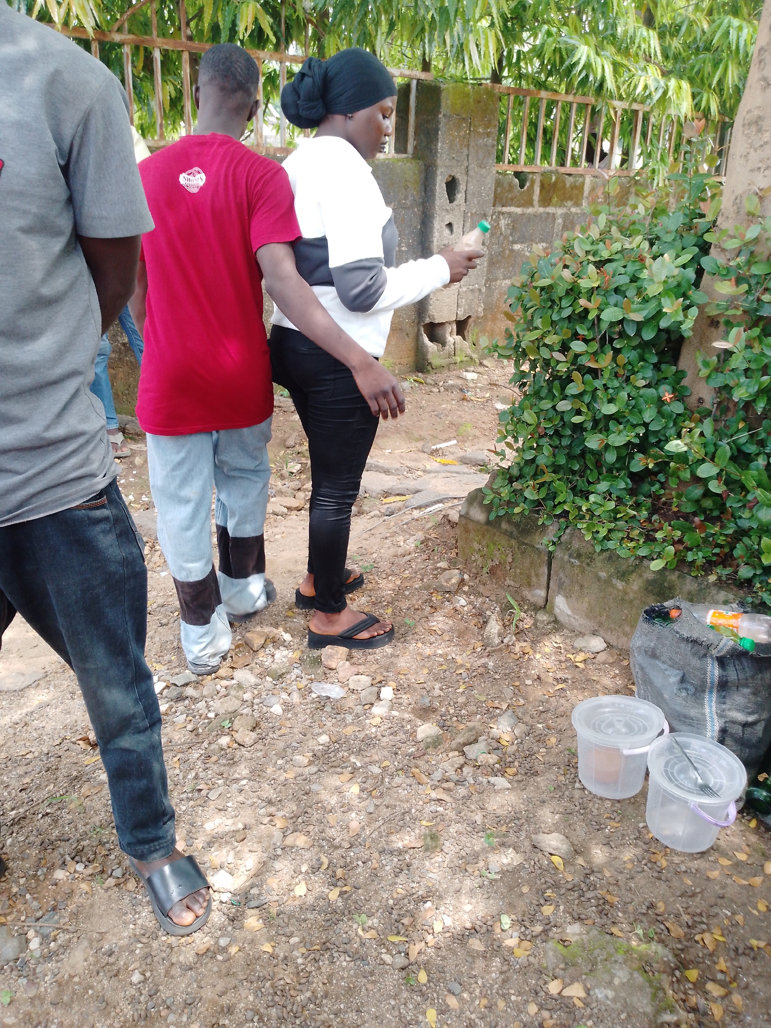 In Akwanga, Nigeria, three individuals are seen standing outdoors on a dirt ground strewn with pebbles and fallen leaves, next to a concrete wall and a large tree. On the left, a person wearing a gray t-shirt and dark blue jeans stands with their back partially to the camera. In the center, a person in a red t-shirt and light blue jeans with dark patches near the ankles is observed from behind. To the right, a person wearing a white long-sleeved shirt with gray accents and black leggings, and a black headscarf, is holding a clear plastic bottle with a green cap. They are looking down at the bottle. To the right of the individuals, a dense green bush with small red flowers grows near the tree. On the ground near the bush, there are two clear plastic buckets with purple handles and a black bag containing items, including a plastic bottle with an orange label. The scene suggests a casual outdoor gathering or a stop along a path.