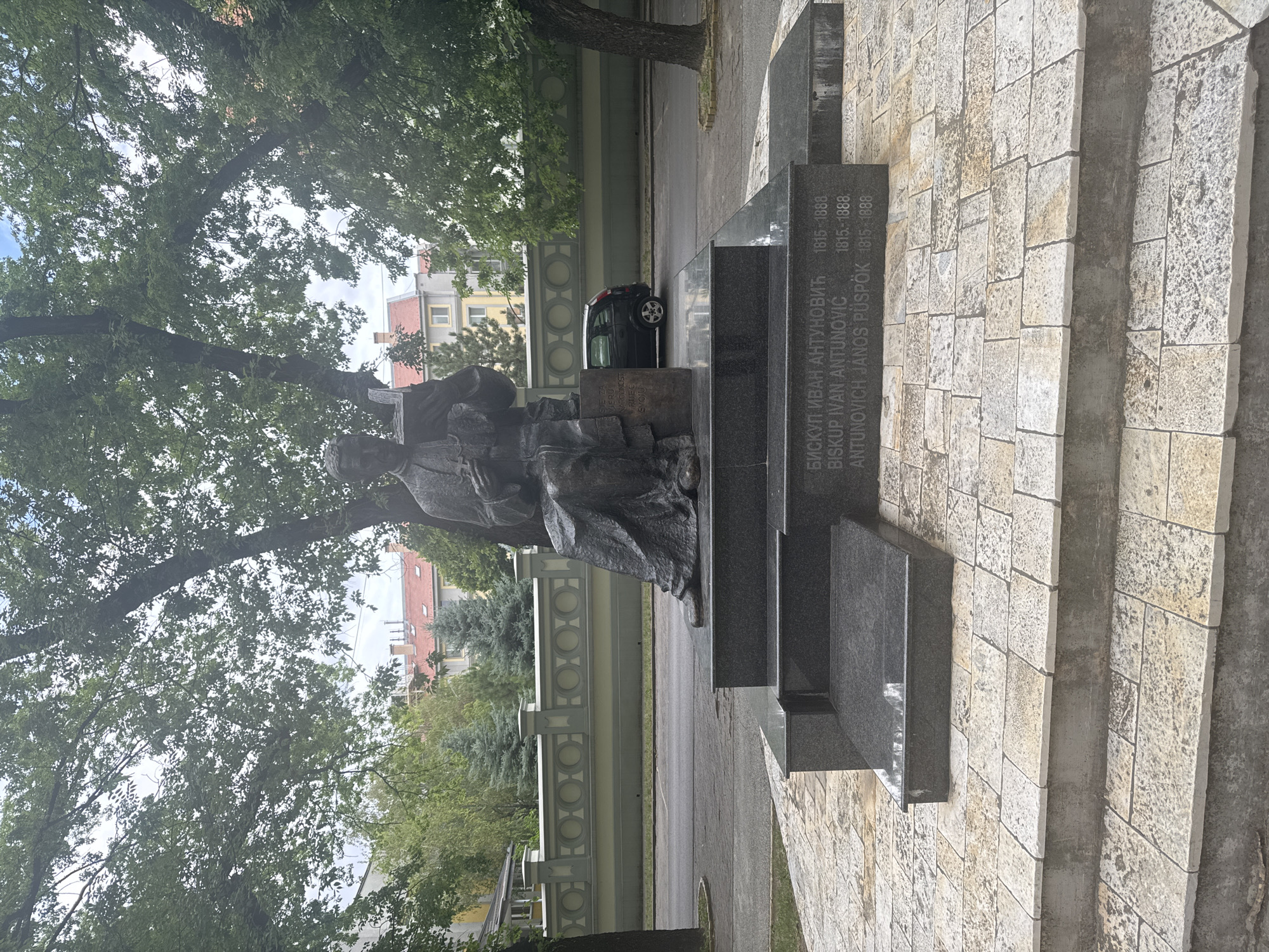 The image shows a bronze statue of a seated man holding a book, likely a historical or religious figure, situated on a granite monument. The monument bears inscriptions in Cyrillic and Latin scripts, along with dates that read "1815-1888". Behind the monument is a street with a black car parked on the side and a decorative fence with circular motifs. Beyond the fence are buildings with red roofs and trees. The statue is located in Subotica, Serbia, specifically in front of the Bishop's Palace, which is part of the Cathedral of Saint Teresa of Avila complex. The statue commemorates Bishop Ivan Antunović, a prominent figure in the Bunjevac community and the Catholic Church in Vojvodina.