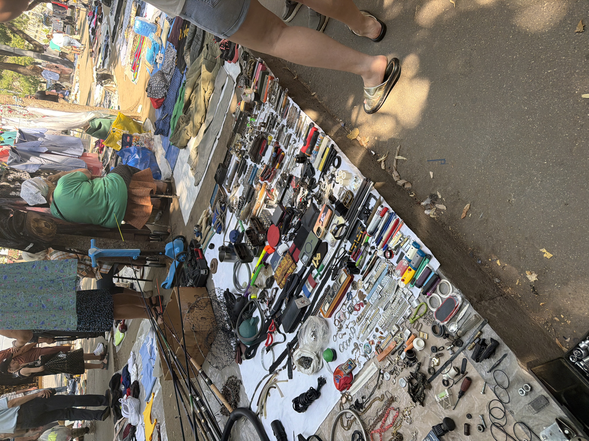 The image depicts an outdoor market scene, likely a flea market or street sale, in Chișinău, Moldova. In the foreground, a table or stall is laden with an assortment of goods, including numerous fishing rods, tools, watches, jewelry, and various small trinkets. The items are spread out on white cloths, with some displayed in boxes or laid directly on the ground. Several people are visible in the background, browsing or tending to the stalls. The ground is paved with asphalt, and sunlight casts shadows, suggesting it is daytime. The overall impression is that of a busy, informal marketplace.