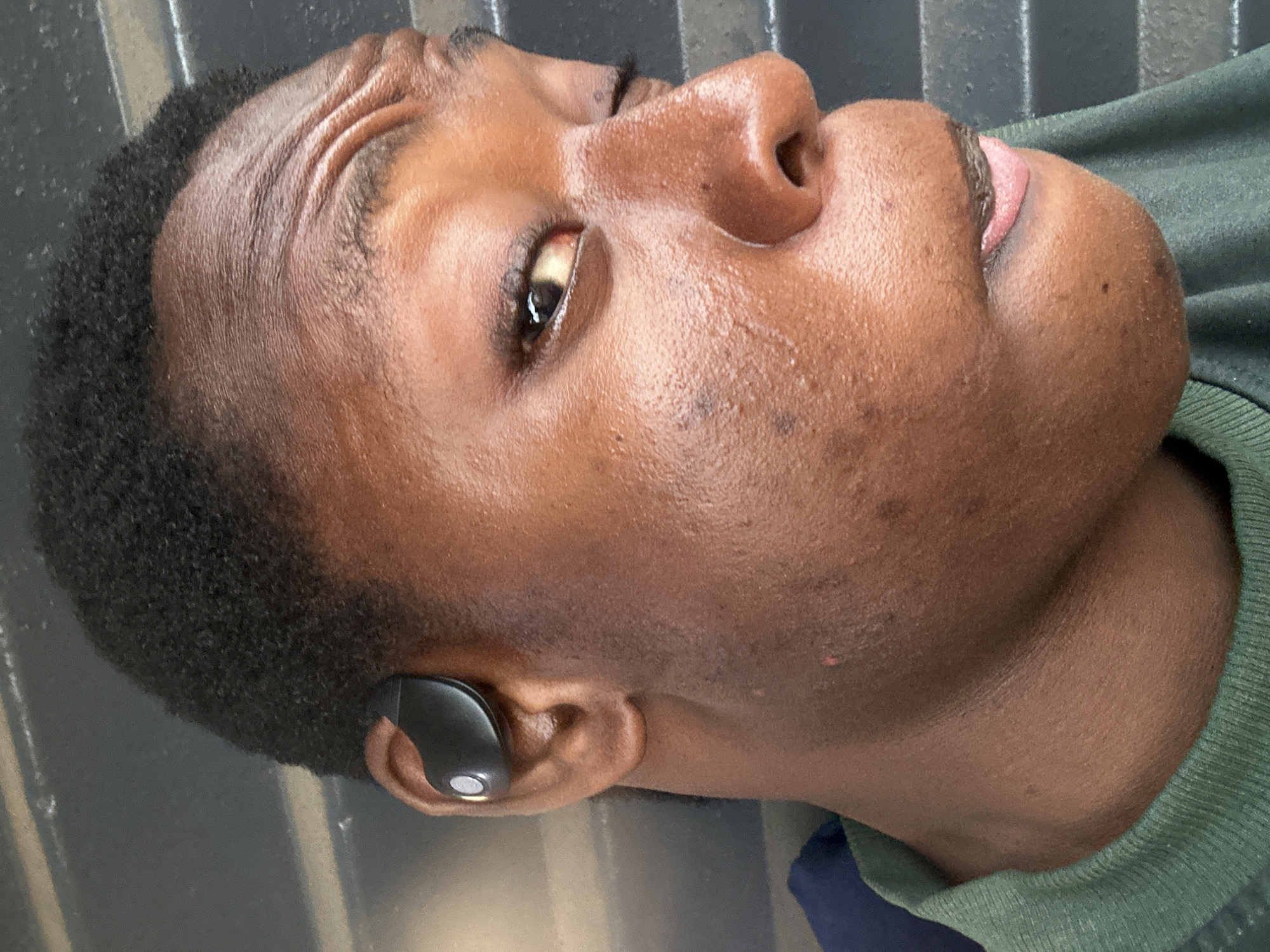 A close-up portrait of a Black male wearing a black wireless earbud in his left ear. The earbud is visible in the ear. The man has short, curly hair. His skin is textured, and there are some visible pores and blemishes. He's wearing what appears to be a green sweater or shirt, with a glimpse of a blue garment underneath. The background appears to be a gray or black surface with some linear patterns. The city and state are unknown.