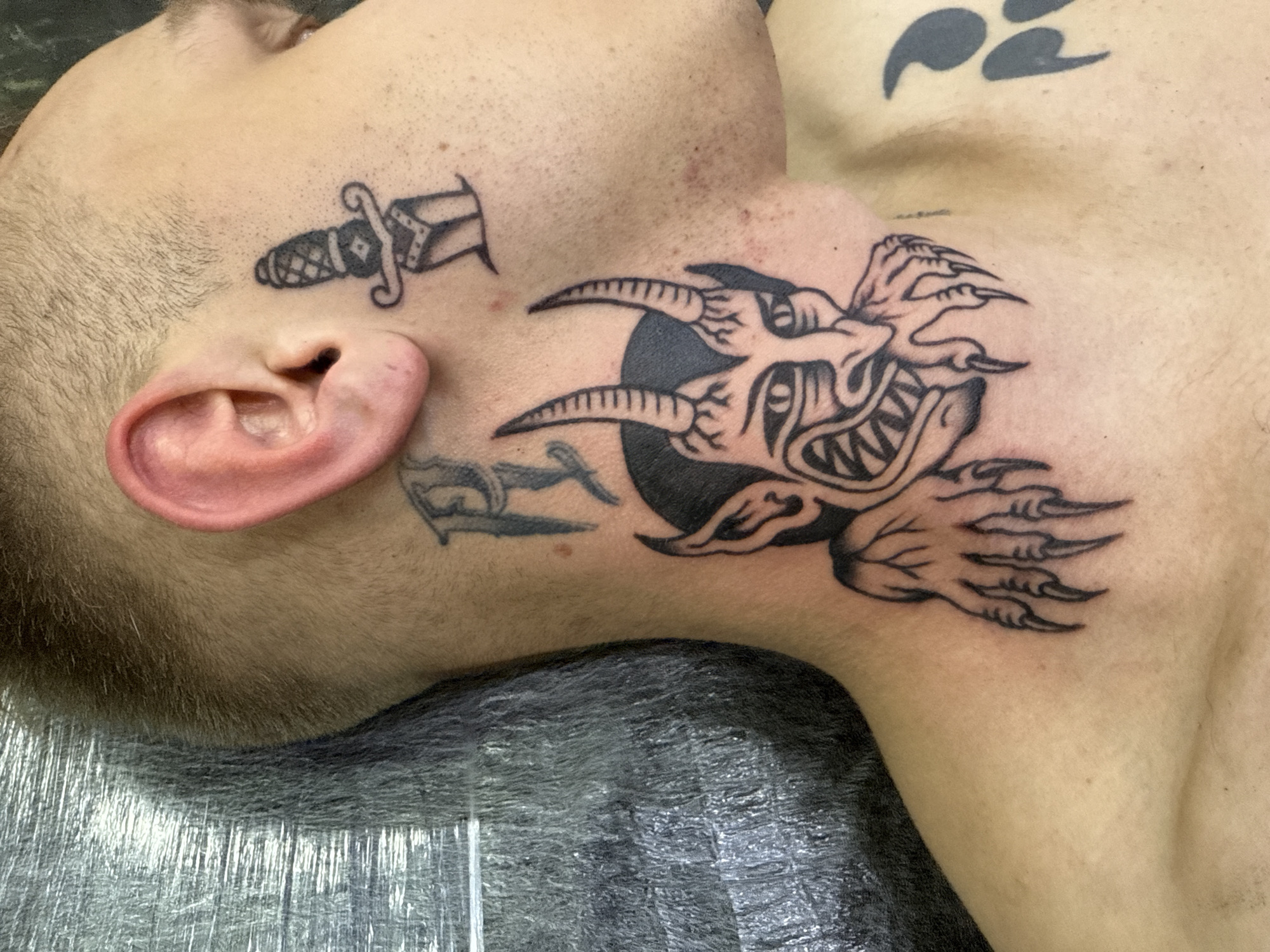A close-up shot depicts a man's face and upper torso, revealing multiple tattoos. On his face, near the ear, a dagger tattoo is visible, with another abstract tattoo located on the lower jawline. A large tattoo resembling a devilish creature is placed on his neck. On his upper chest is another tattoo consisting of three claw-like shapes. The man is lying down on a surface covered in a shiny, plastic-like material. The background is out of focus. There is no evidence indicating this image was taken in Moscow, Russia.