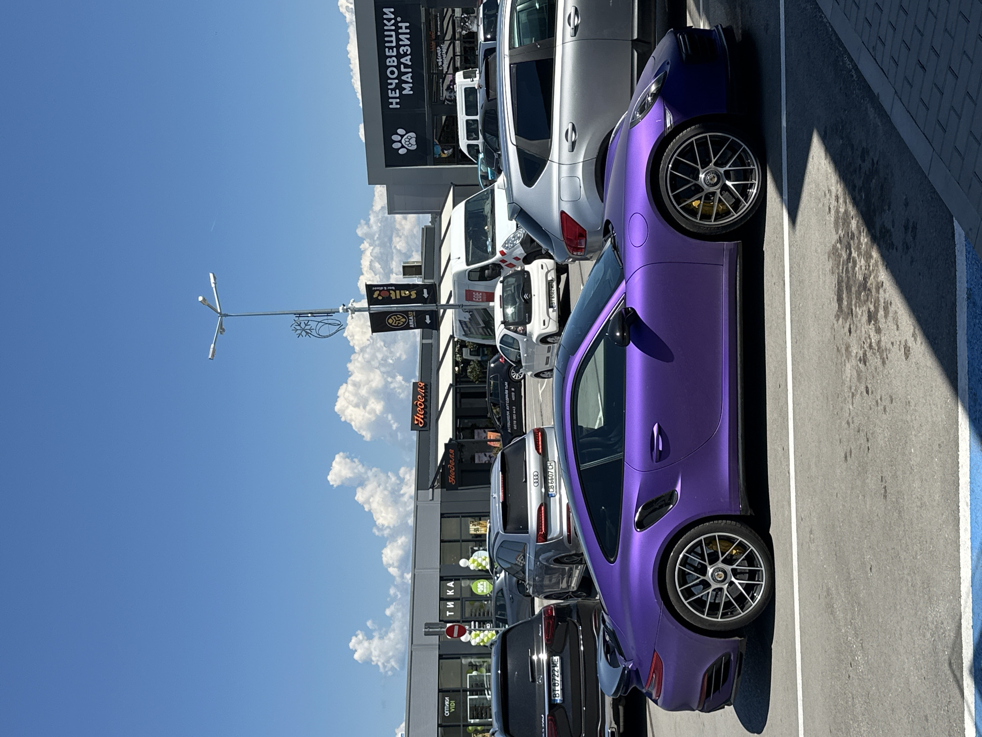 A bright, sunny day in Veliko Tarnovo, Bulgaria, captures a parking lot outside a commercial building complex. The sky is a vivid blue with scattered fluffy white clouds. In the foreground, a striking matte purple Porsche sports car is parked. Behind it, a black Mercedes SUV and a silver Audi SUV are visible. Further back, several other vehicles, including a white van and a small white car, are parked. The building in the background features storefronts with signage, including "Нечовешки Магазин" (meaning "Inhuman Store") with a paw print logo, and "Тикa" (meaning "Tick-tock"). The architecture is modern, with a gray facade and a black roof. Streetlights and advertising banners are also present in the scene. The overall impression is a typical commercial area on a clear day.