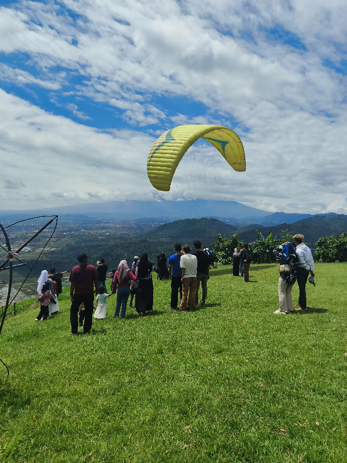 The image shows a group of people gathered on a grassy hillside overlooking a valley in Majalengka, Indonesia. A bright yellow and green paraglider is flying overhead, its lines connected to a launch point on the hillside. The group is comprised of at least 15 people, including men, women, and children. The individuals are all wearing casual clothing. They are watching the paraglider as it ascends. The sky is clear with white, puffy clouds scattered across it. In the distance, the valley is shrouded in haze and appears to be populated with small buildings and trees. The environment is lush and green, with rolling hills and trees in the background. The hillside is covered with short, green grass.