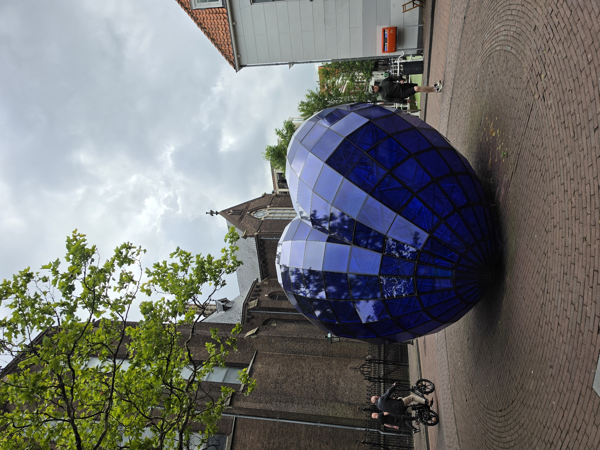 The image depicts a street scene in Delft, Netherlands, featuring a large, translucent blue heart-shaped sculpture prominently in the foreground. The sculpture is composed of geometric panels and reflects the cloudy sky and surrounding buildings. To the left of the sculpture, a brick building with arched windows, possibly a church, is visible, partially obscured by the branches of a leafy tree. In front of the brick building, a black metal fence lines the sidewalk.
Two individuals are present. One person is riding a black electric bicycle, wearing a dark jacket and beige pants, and is positioned to the left of the heart sculpture. Another person, wearing a dark jacket, shorts, and a cap, is walking on the sidewalk to the right of the sculpture.
The ground is paved with red bricks arranged in a circular pattern around the sculpture. In the background, a white building with an orange-tiled roof is visible on the right side of the image. An orange mailbox is mounted on the wall of this building. Trees and more buildings are visible further in the background. The sky is overcast with a majority of gray clouds.